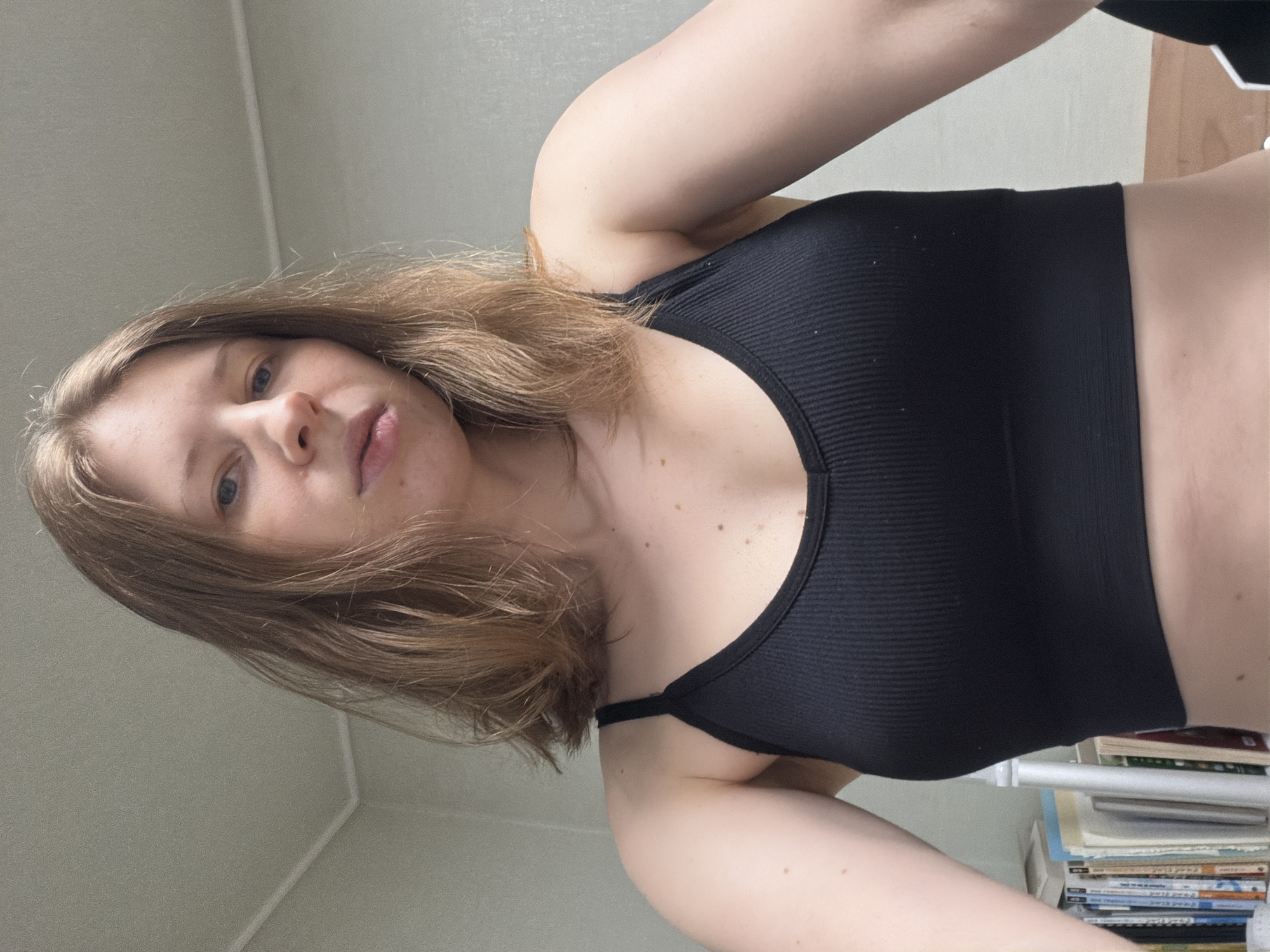 A young woman with shoulder-length brown hair is captured in a low-angle, upward shot. She is wearing a black ribbed sports bra. Her face is turned towards the camera, and she appears to be looking directly at it. Her eyes are blue, and her lips are slightly parted. The background consists of a plain, light-colored wall, with the corner of a bookshelf visible on the right side of the frame. The bookshelf is filled with various books, some of which have visible text, though it is not legible. The lighting suggests it is daytime, and there are no clear indicators of the weather or specific time of day beyond that. The overall mood is casual and intimate, possibly a selfie taken in a private setting like a bedroom or living area. The location context is Dongducheon-si, South Korea.