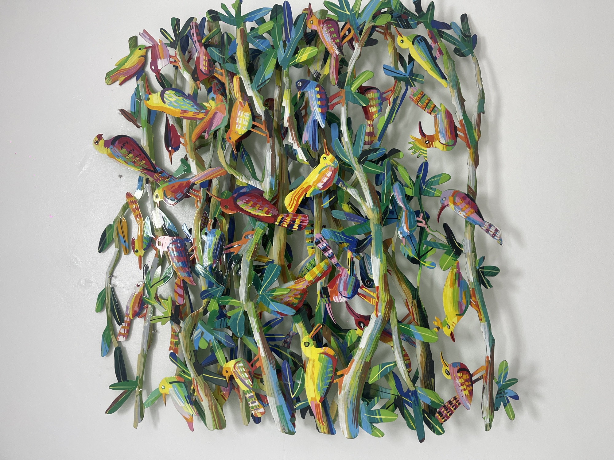 A colorful, multi-layered artwork depicting birds and trees is mounted on a white wall. The artwork is comprised of numerous individual components, likely made of a thin material such as paper or cardstock, and painted in bright, varied colors including blues, greens, yellows, reds, and browns. The birds are depicted in various poses, perched on the trees, with a high level of detail in their feathers and features. The trees are simplified, vertical forms with leaves extending outwards. The artwork has a three-dimensional quality due to the layering of the elements. The background is a plain, white wall. Location: Unknown, Unknown.