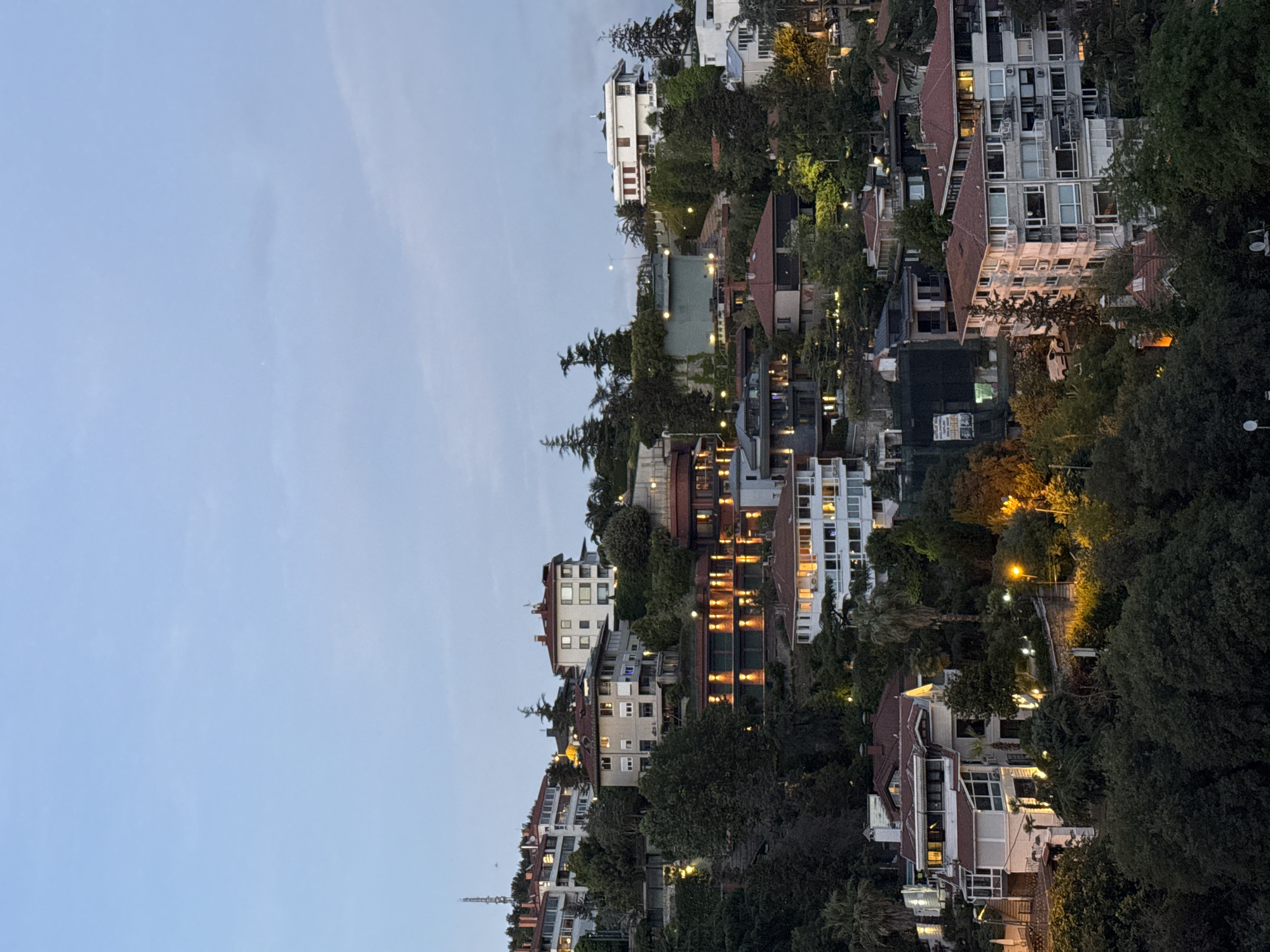 The image depicts a hillside residential area in Beşiktaş, Turkey, as twilight descends. The scene is characterized by a cluster of multi-story buildings nestled among lush green trees. Many of the buildings are illuminated from within, suggesting the evening hour and the start of artificial lighting. The architecture varies, with some structures featuring modern designs and large windows, while others exhibit more traditional tiled roofs.
The sky is a soft, pale blue, indicating it is either early evening or late afternoon, transitioning towards night. The lighting in the scene is a mix of natural twilight and the warm glow emanating from the windows of the houses and from streetlights scattered throughout the area, illuminating pathways and trees. The dense foliage suggests a natural or well-landscaped environment integrated with the urban development.
The perspective is from an elevated viewpoint, looking down and across the hillside, giving a sense of depth and the layered nature of the community. There are no visible people, but the lit windows imply the presence of residents. The overall mood is tranquil and serene, capturing a quiet moment in a densely populated area. A tall tower, possibly a communication mast or a landmark, is visible in the distance on the left side of the frame. The image does not contain any discernible text.