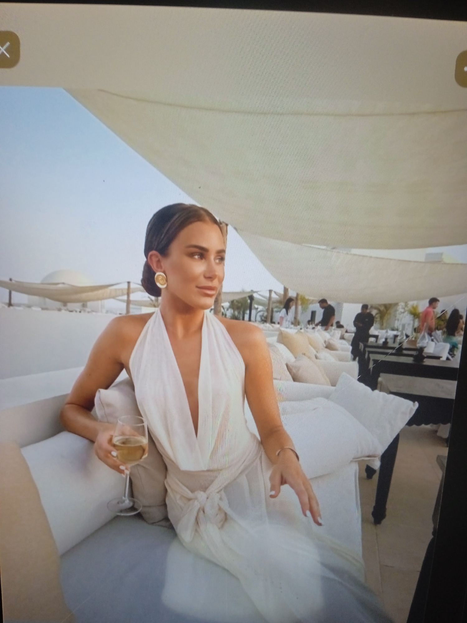 A woman is seated on a cushioned sofa outdoors, holding a glass of champagne. She is wearing a white halterneck dress and large gold earrings. The background shows people dining at tables under a white canopy. The location is not Chernivtsi, Ukraine.