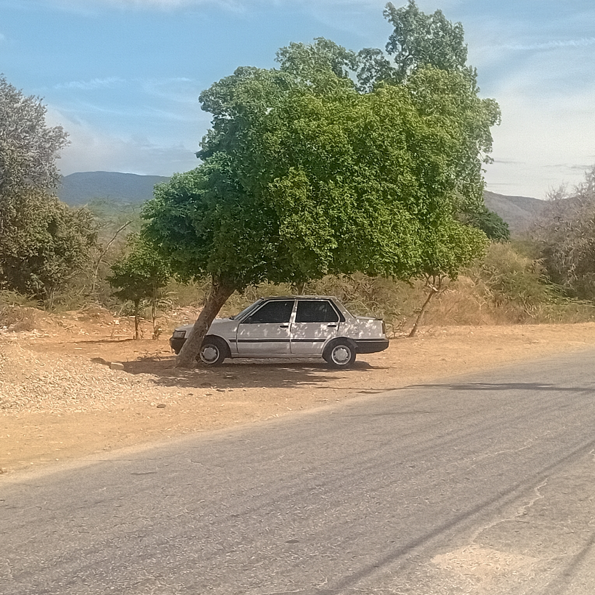 A silver four-door sedan is parked under the shade of a large, leafy tree beside a paved road. The surrounding environment is dry and dusty, with sparse vegetation and small hills in the background. A pile of light-colored debris is visible to the left of the car. The sky is partly cloudy. The location is Unknown, Unknown.
