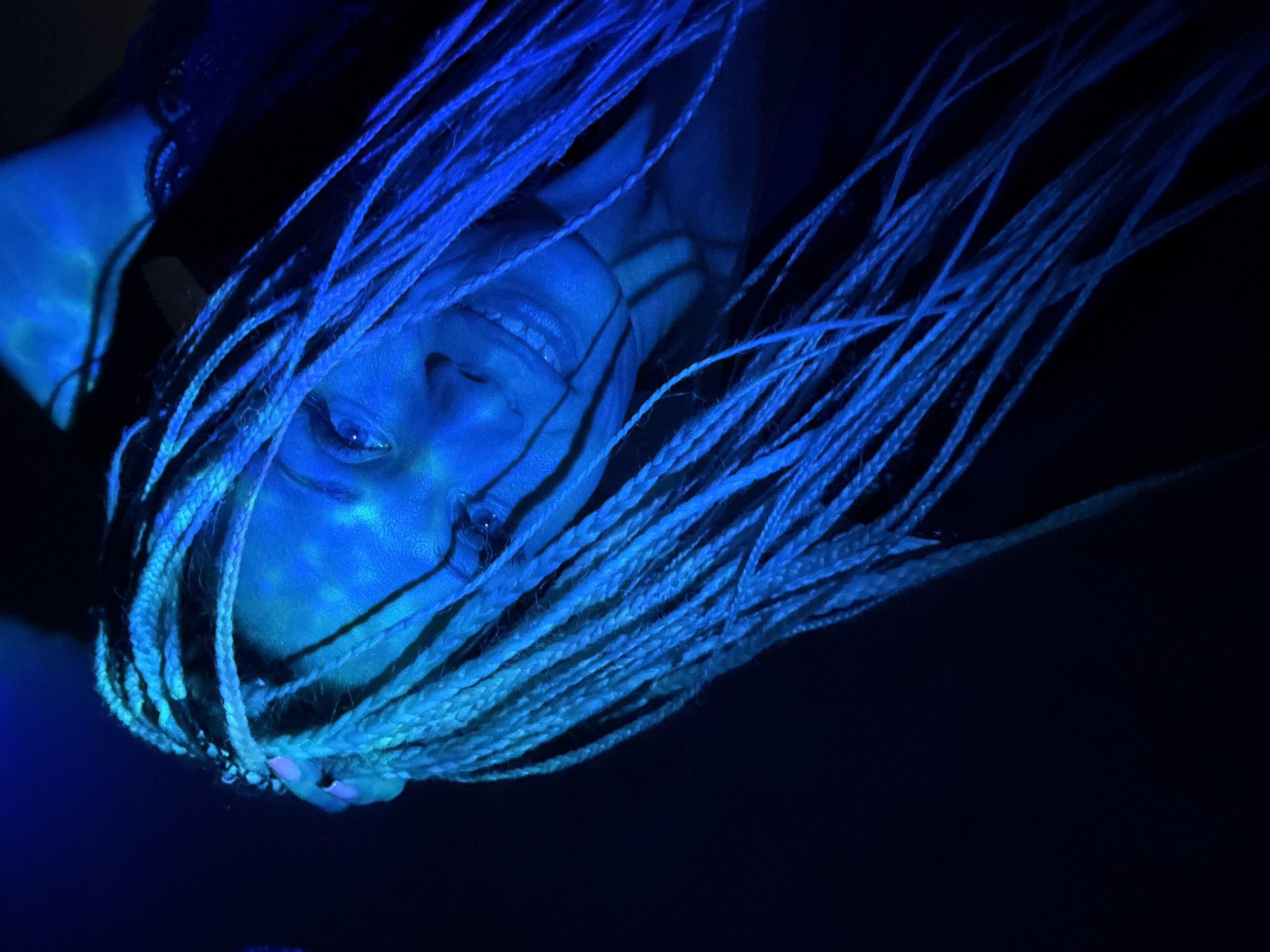 In a dimly lit room, a person with blonde braided hair is looking upwards, their face illuminated by blue light. Their eyes are open, and they are smiling, revealing their teeth. The background is dark and indistinct. No specific location in Zhukovo, Russia, can be determined from the image.