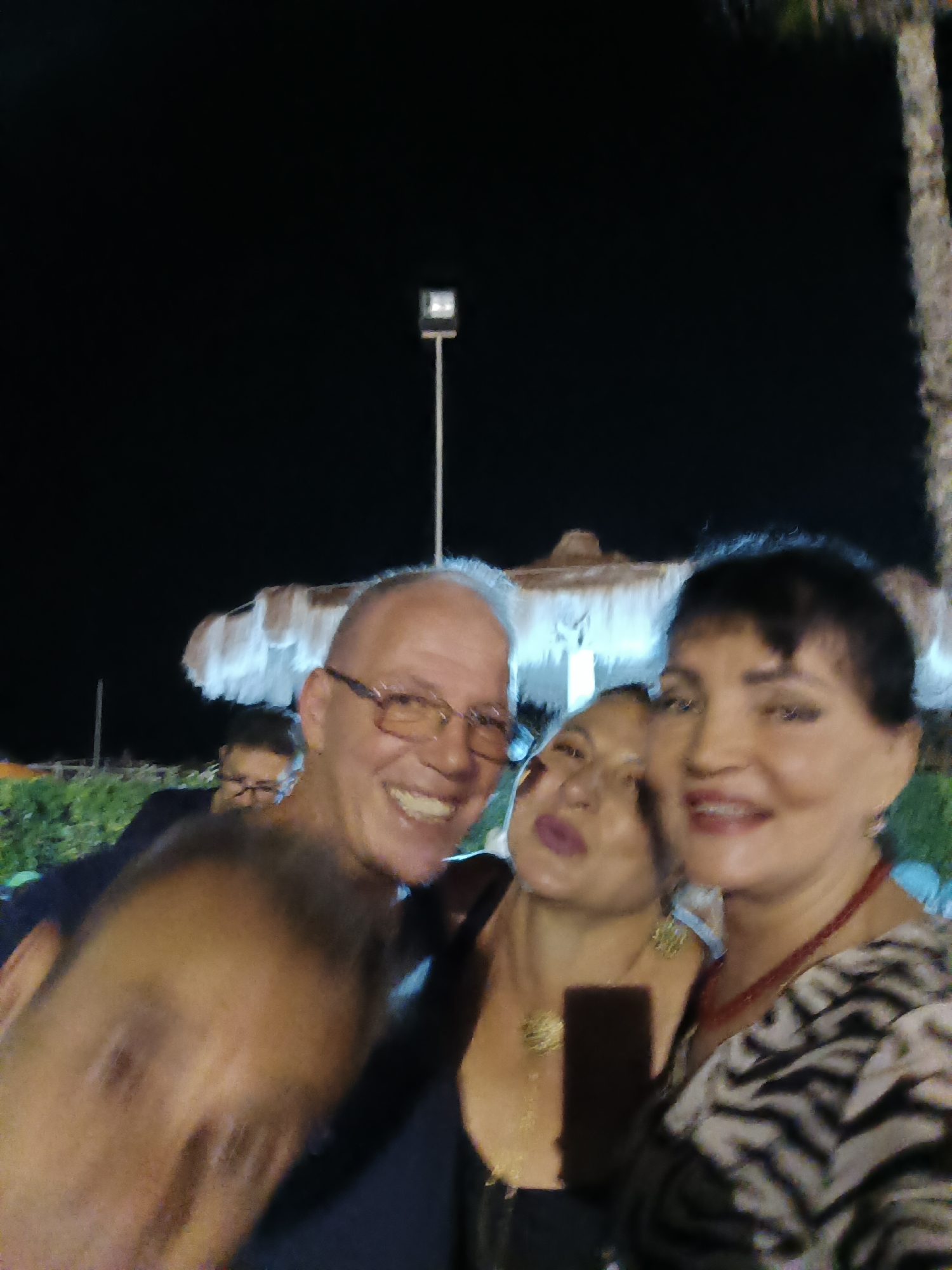 Four individuals are present in the image. In the foreground, a man wearing glasses is smiling, with his right arm partially visible. Next to him, a woman with dark hair and a black top is smiling, and a second woman, also with dark hair and wearing a black top with a gold necklace, is positioned behind them and looking upwards. A fourth person, a man wearing glasses, is visible in the background, to the left of the three individuals in the foreground. The scene appears to be outdoors at night, with artificial lighting evident from a street lamp and possibly other light sources illuminating umbrellas and foliage in the background. The overall setting suggests a social gathering in a public area.