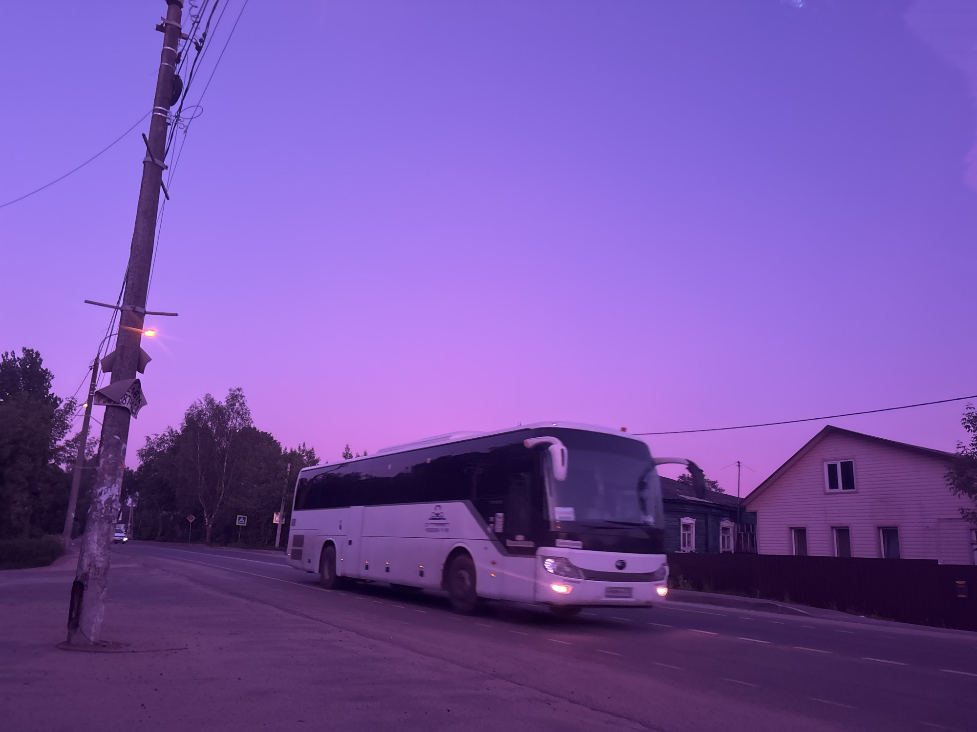 A white bus drives along a road in Zaprudnya, Russia. To the left, a weathered utility pole with wires and a streetlight stands next to a sidewalk. Behind the bus, a wooden house is visible next to a dark fence. Trees line the background, and the sky is a uniform purple.