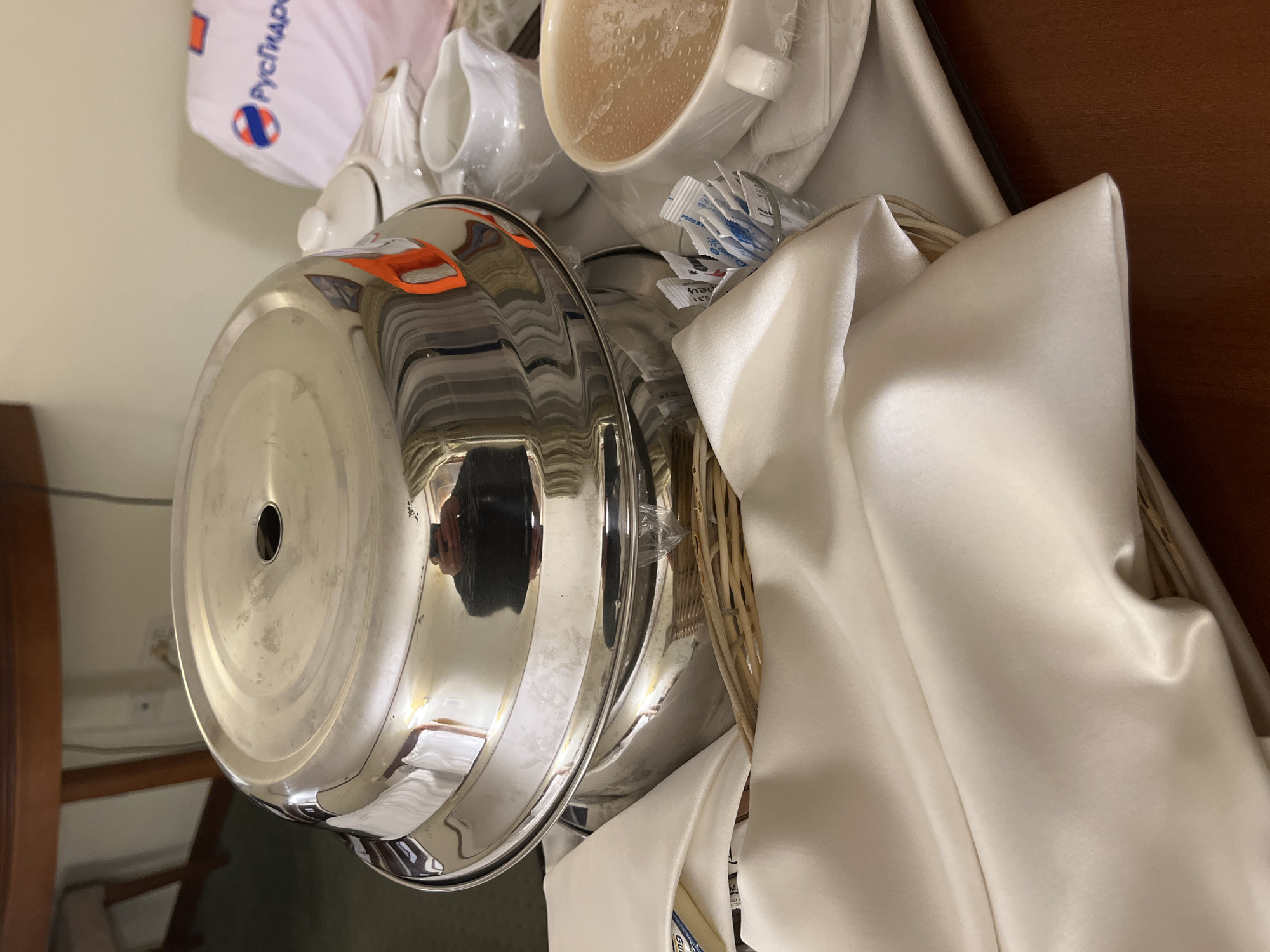 A close-up, rotated image shows a hotel room service tray. The tray is covered with a white cloth and contains a cup of soup, creamers, sugar packets, and a covered dish. The reflection in the dish cover suggests the photo was taken indoors, possibly in Moscow, Russia, given the presence of a "Русгидро" branded item, a Russian hydroelectric company. The image gives no information about people, interactions, or actions. The environment consists of a typical hotel room.