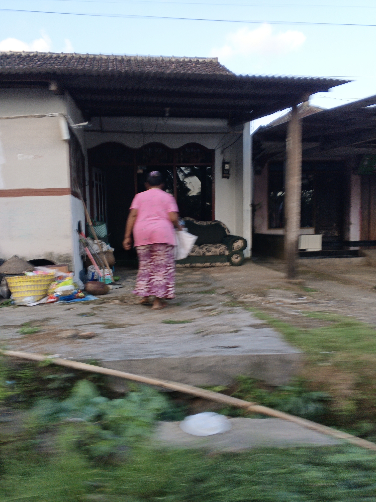 A person wearing a pink shirt and a patterned skirt walks away from the camera, carrying a white bag. The person is walking on a concrete pathway in front of a house with a dark-tiled roof. To the left of the pathway, there is a yellow basket filled with colorful plastic bags and other items. Several brooms and cleaning tools are leaning against the wall of the house. A dark-colored couch with a patterned seat is visible on the porch. The environment appears to be an outdoor residential area in Balak, Indonesia, with green vegetation and some dirt or gravel on the ground. The image has a blurred effect, suggesting it was taken while in motion.