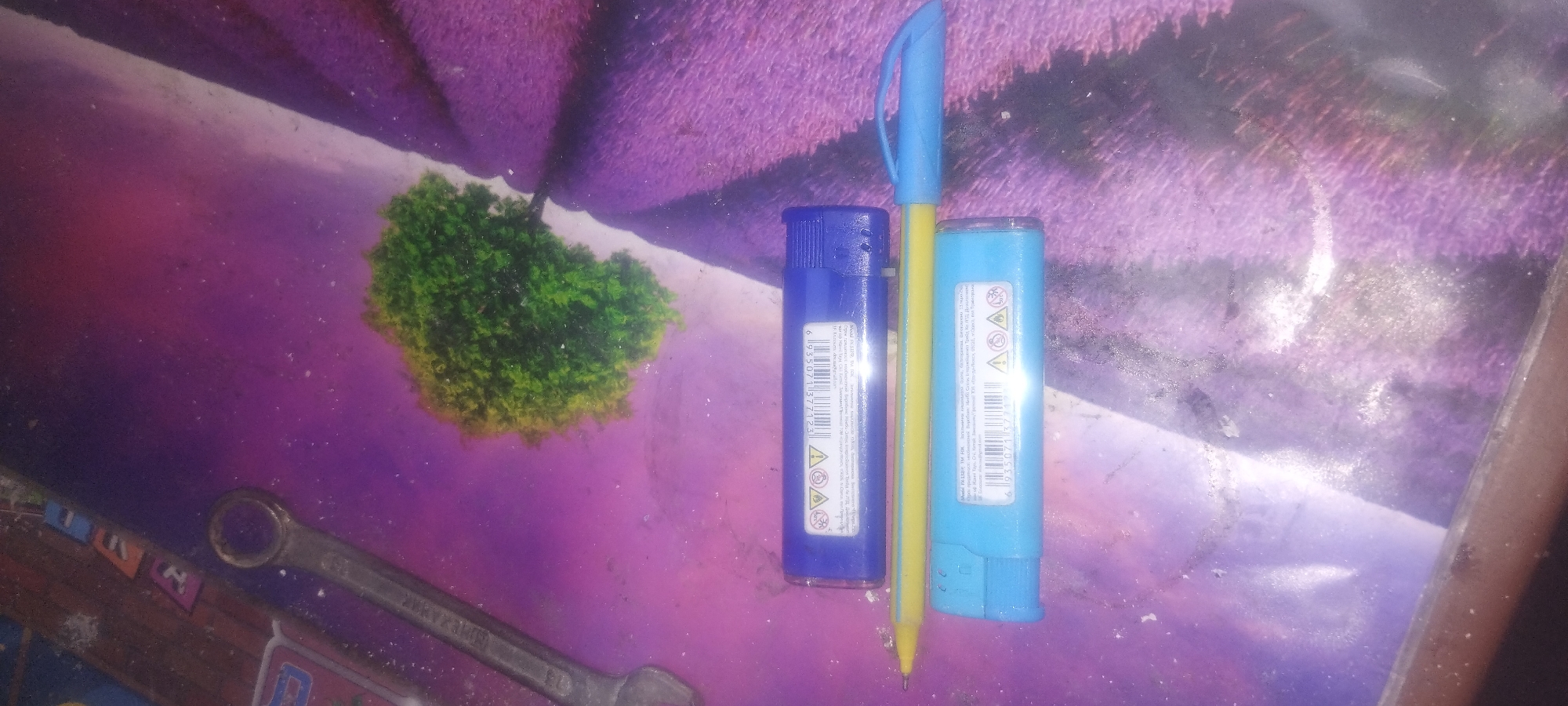 An overhead, slightly angled shot shows a blue and a lighter blue object, likely lighters, on a purple patterned surface. A yellow pen is situated between the two objects. To the left, a silver wrench is visible. In the background, a small, green, artificial tree sits on the same purple surface. The surface appears to be a placemat or table covering with a digitally printed image of a tree and a purple, textured background. The scene is lit from above, casting subtle shadows. The objects are stationary. There are no people present. The location is unknown.