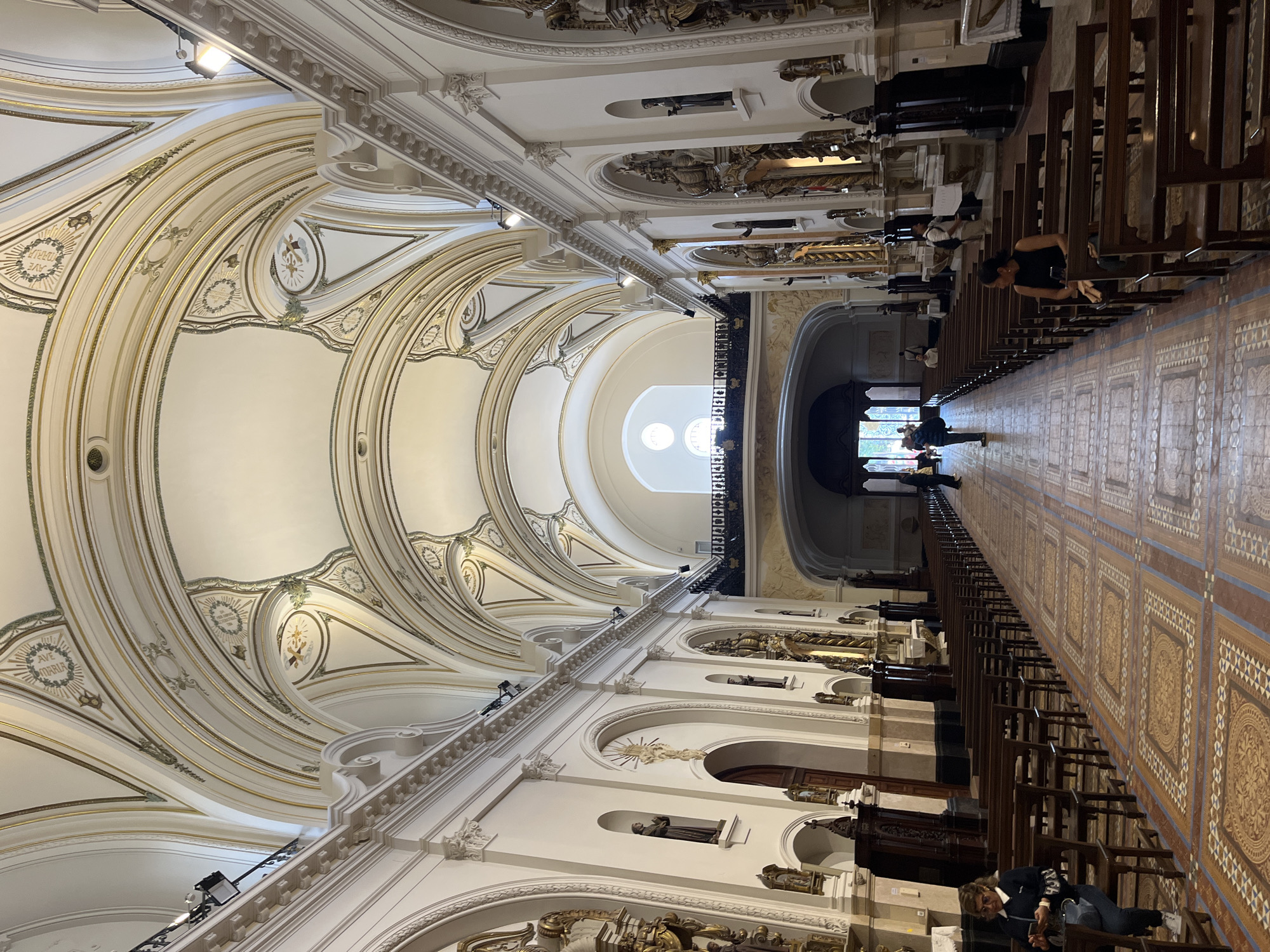 This image depicts the ornate interior of a grand church or cathedral, likely located in Buenos Aires, Argentina. The perspective looks down the length of a long central aisle, showcasing the impressive architecture from a slightly elevated vantage point.
The most striking feature is the high, vaulted ceiling, adorned with intricate white and gold/cream-colored decorative plasterwork. Numerous arches, ribs, and medallions embellish the ceiling, with spotlights strategically placed to highlight their details. One medallion clearly displays the Latin inscription "AVE MARIA."
Rows of dark wooden pews line both sides of the aisle, extending into the distance. The aisle itself features a decorative patterned floor, possibly of tile or inlaid wood, with geometric and curvilinear designs in shades of brown, tan, and white.
Several people are present in the church. On the right side, a woman with dark hair is seated in a pew, looking down. Further along the aisle, other individuals are visible standing or seated, suggesting they are visitors or congregants. Towards the far end, near what appears to be an exit or another section of the church, more people can be seen.
The side walls are adorned with architectural elements such as pilasters, arches, and niches containing religious statues, consistent with a place of worship. An upper gallery or triforium level with a black balustrade is also visible. The overall impression is one of grandeur and reverence, with natural light entering from windows higher up, indicating it is daytime. The architecture suggests a Baroque or Neoclassical influence typical of historical religious buildings in the region.