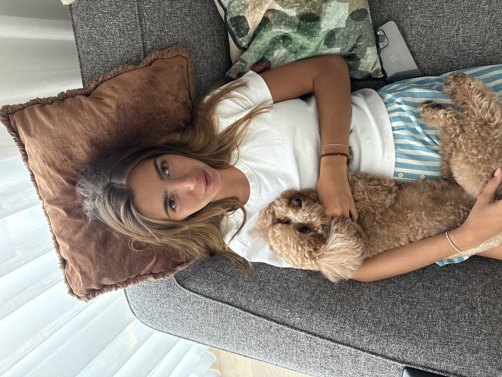 A young woman with long, light brown hair lies on a grey sofa, holding a small, fluffy, light brown poodle. She is wearing a white t-shirt and blue and white striped pajama bottoms. A brown throw pillow with fringe is behind her head, and a green and brown camouflage patterned pillow is to her right. A smartphone is visible under the camouflage pillow. The scene appears to be an indoor setting, likely a living room, with natural light coming from a window with white blinds. The setting is not specifically identifiable as being in Loures, Portugal.