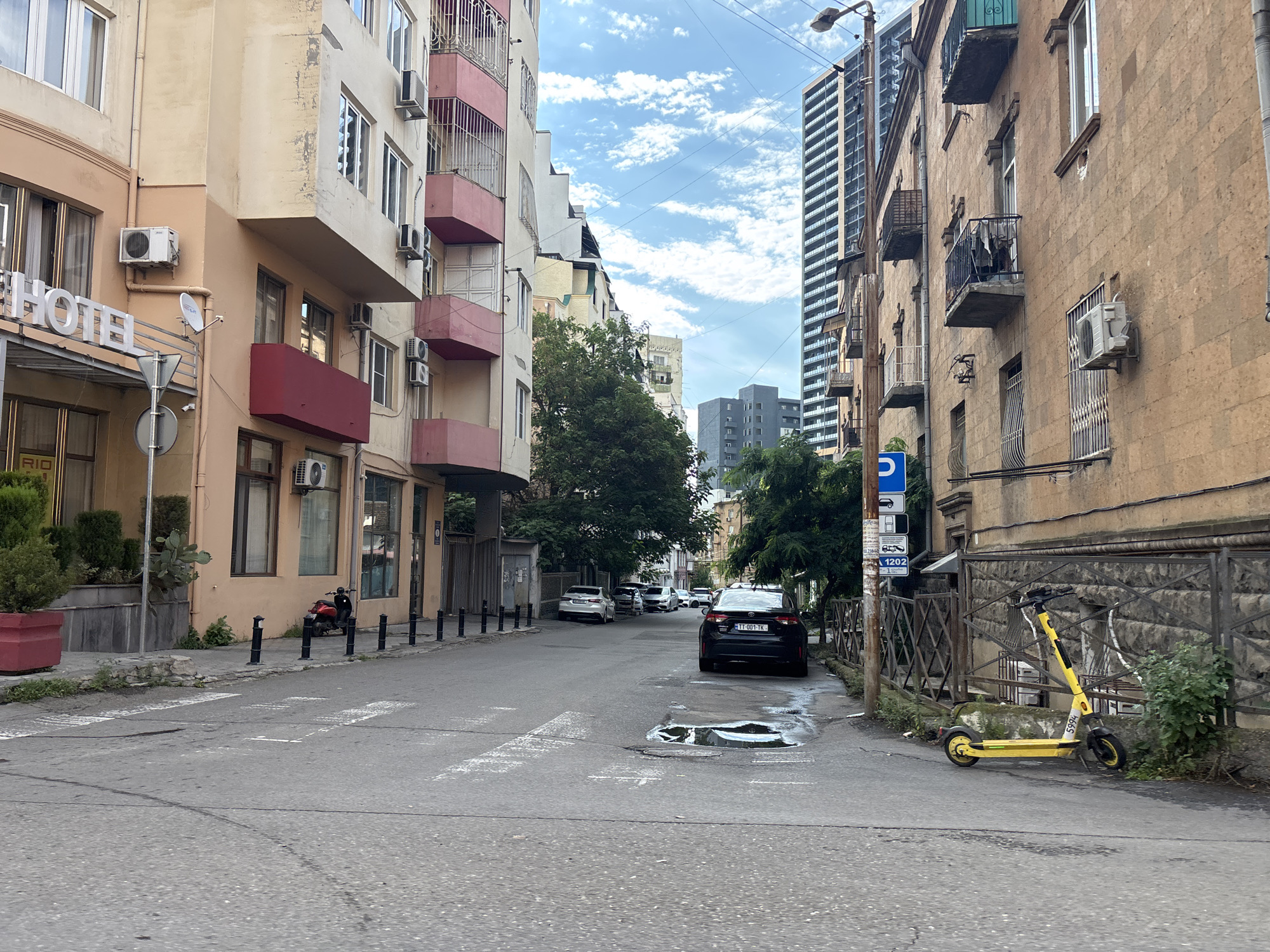 The image depicts a street scene in Tbilisi, Georgia, likely in a residential or mixed-use area. The street is paved and shows signs of wear, including a large puddle near a pedestrian crossing. On the left side of the street, a multi-story building with a light peach facade is visible, featuring multiple windows, air conditioning units, and a prominent "HOTEL" sign. Red balconies with metal railings adorn this building.
On the right side, another building with a rough, textured facade in a similar peach hue is present. This building also has windows and balconies, some with laundry hanging. A dark-colored car with a license plate reading "TT 001-TK" is parked on the right side of the street, partially obscuring a metal fence. Beyond the car, a yellow electric scooter is parked on the sidewalk, leaning against the fence.
Several other cars are parked or driving on the street, including a white sedan further down on the left and a black sedan in the distance. The street is lined with black bollards along the sidewalk. Trees are visible in the background, with a tall modern skyscraper also present in the cityscape. The sky is partly cloudy. A red scooter is parked on the sidewalk on the left, near a planter.