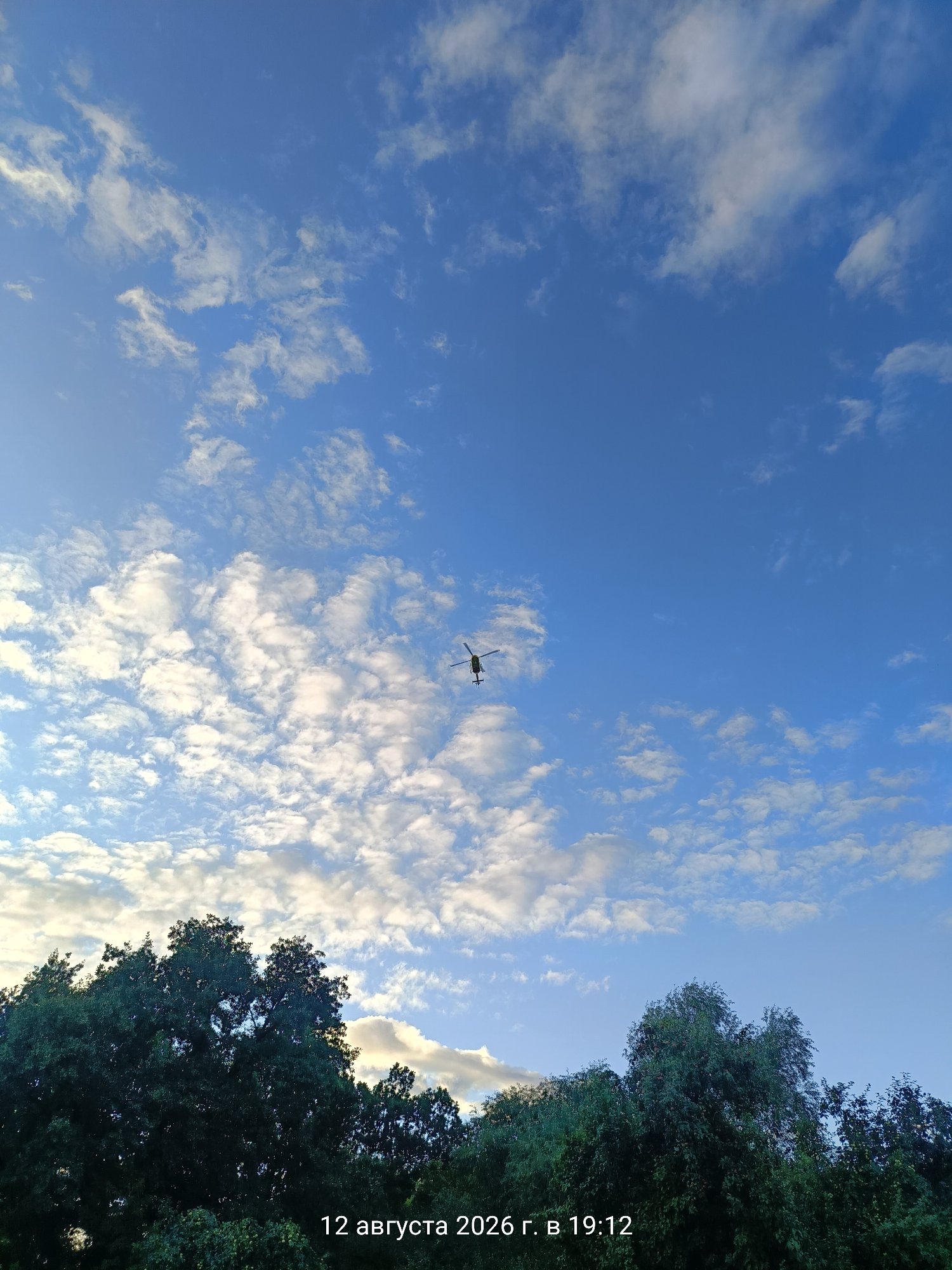 A low-angle shot shows a clear blue sky filled with scattered white clouds. In the upper center of the frame, a dark-colored helicopter is seen in mid-flight, its rotors spinning. The lower portion of the image is dominated by the dark green foliage of trees, obscuring the ground. A date and time stamp, "12 августа 2026 г. в 19:12", is visible in the bottom right corner.