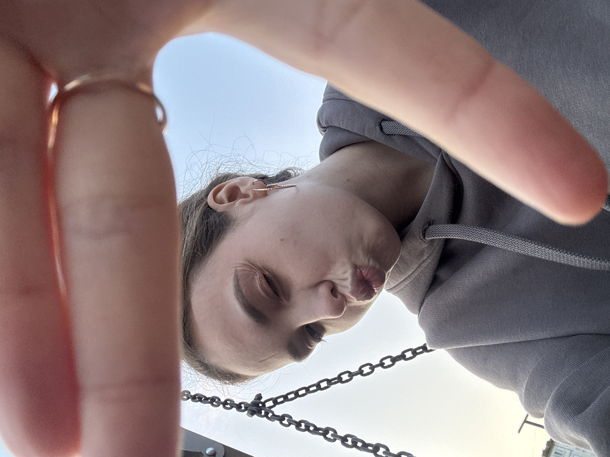 The image is a close-up, selfie-style shot of a young woman with light brown hair, wearing a grey hooded sweatshirt. She is making a kissing face with her eyes partially closed. A gold ring is visible on her right index finger, which is held up close to the camera, partially obscuring the shot. A chain, possibly from a swing, is visible on the left side of the frame, and the sky is visible in the background. The lower portion of the image shows the woman's shoulder and arm, also partially covered by her hand. There is no discernible indication of the location being in Noviy, Russia.
