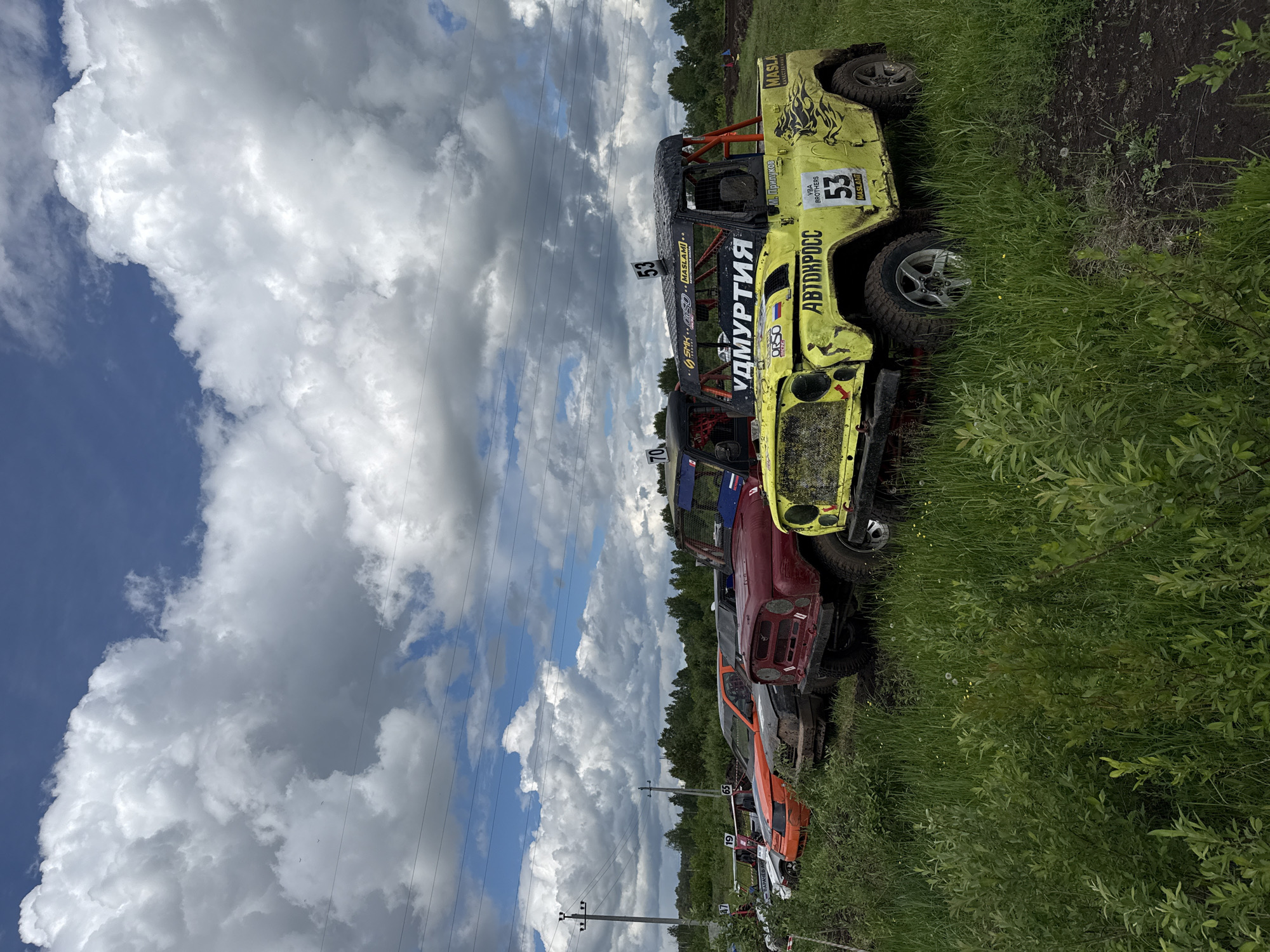 The image shows a line of off-road racing vehicles parked on a grassy field. The foreground vehicle is a lime green off-road vehicle with the number "53" and the words "Удмуртия" and "Автокросс" in Cyrillic script. Several other vehicles are parked behind it, including red, orange, and white ones. Some have racing numbers such as "19", "65" and "70". The background features a cloudy sky and a line of trees. Power lines can be seen stretching across the sky. The location is likely near Votkinsk, Russia, based on the Cyrillic text and implied setting of off-road racing.