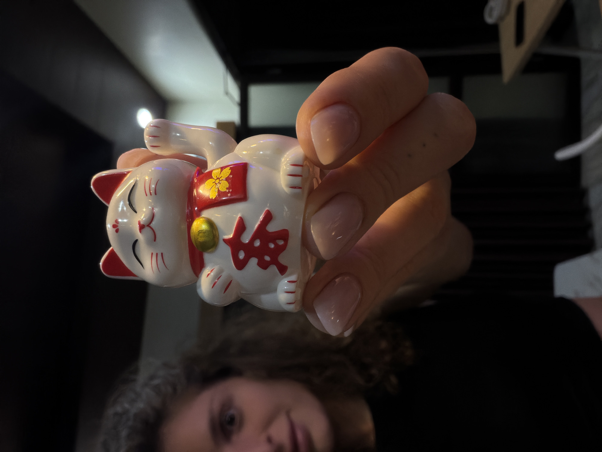 A close-up, portrait-oriented shot captures a person's hand holding a white Maneki-neko (lucky cat) figurine against a dark, out-of-focus background.
The Maneki-neko is primarily white with prominent red detailing. Its ears are tipped in red, a red collar encircles its neck with a small golden bell, and its paws have red stripe accents. On its belly, a large red kanji character, "福" (fuku), signifying good fortune or luck, is prominently displayed. Above this character, a red banner features a yellow floral or clover design. The cat's left paw is raised in a traditional beckoning gesture.
The hand holding the figurine belongs to a person whose face is partially visible in the bottom portion of the frame. The person has dark, curly hair and looks towards the camera with a neutral to slightly smiling expression. The hand features neat, light-colored fingernail polish.
The scene appears to be indoors, under low-light conditions, as indicated by the dark and blurry background, which also shows some subtle reflections or light sources. There are no direct visual cues to the location, time of day, or weather beyond an indoor setting.