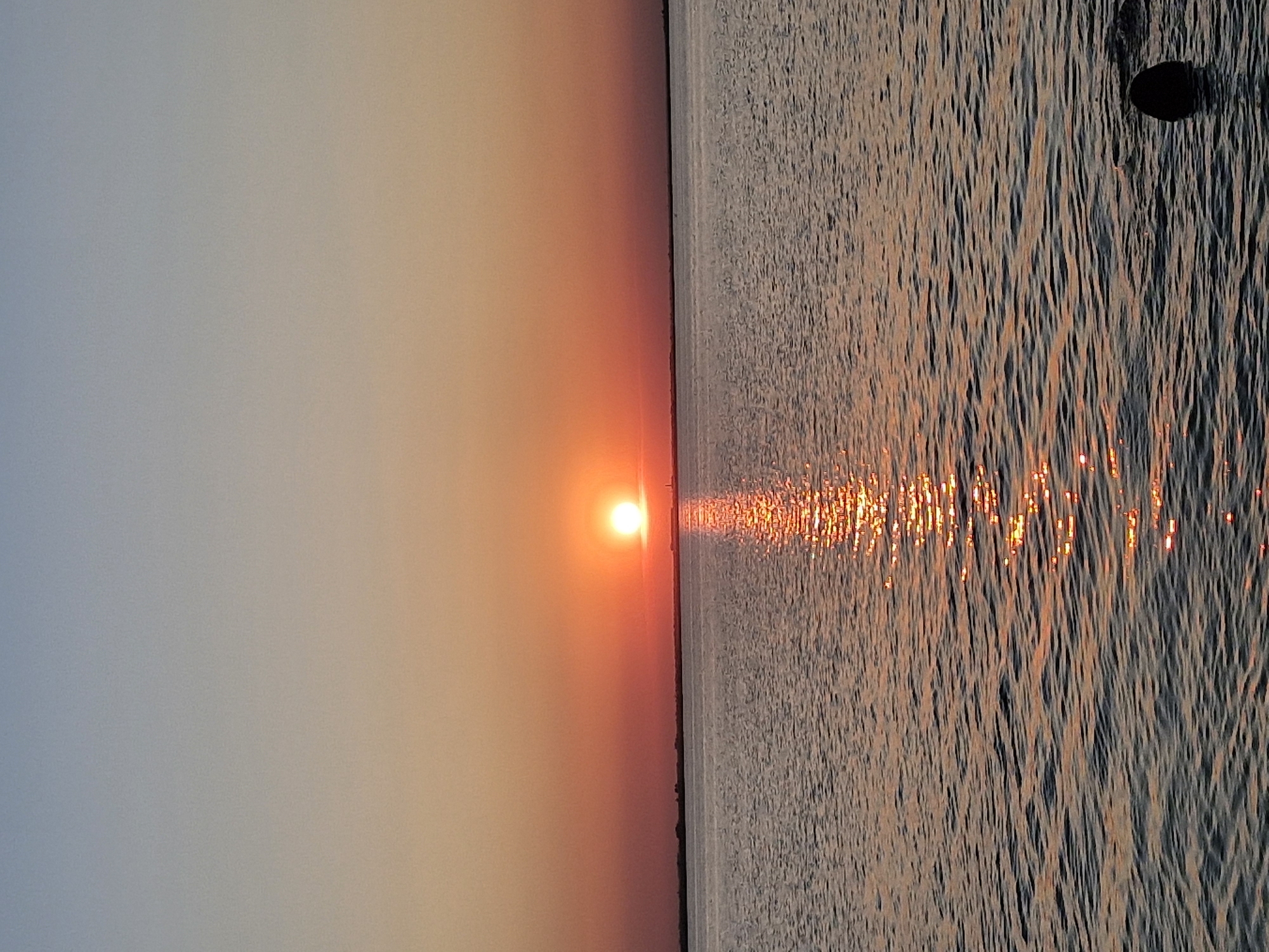 A sunset over a body of water is captured in this vertical, medium-shot photograph. The sun, a bright orange orb, is low on the horizon, casting a shimmering reflection on the rippling water. The sky transitions from a pale blue at the top to a warm orange and red hue near the horizon, blending with the color of the sun. A dark, horizontal line on the horizon suggests land or a distant shoreline. The water in the foreground is dark with subtle waves, and a dark, rounded object, possibly a buoy or a submerged object, is visible on the right side of the frame. The scene is serene and natural, with no discernible human activity or urban elements, making it difficult to pinpoint a specific location within Kokshetau, Kazakhstan, without further context.