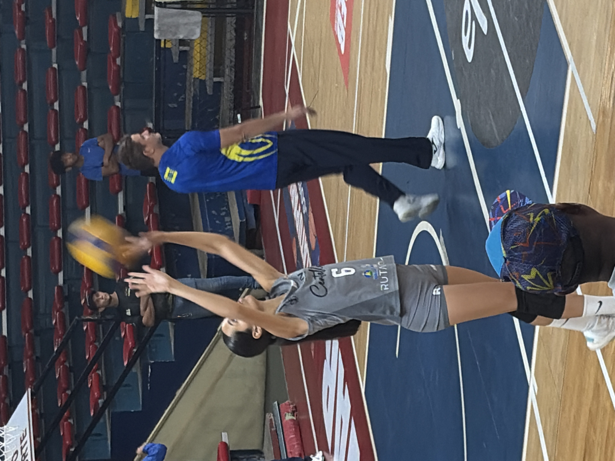 A woman in a gray volleyball uniform with the number 6 on the front is in the act of receiving a volleyball, her arms extended upwards. She is on a basketball court with blue and tan painted areas. In the background, there is a basketball hoop and bleachers. A man in a blue shirt and dark pants, and another man in a blue shirt are also visible on the court. There are additional people in the bleachers. The environment suggests an indoor basketball arena or gymnasium, which could be located in Ciudad Guayana, Venezuela.