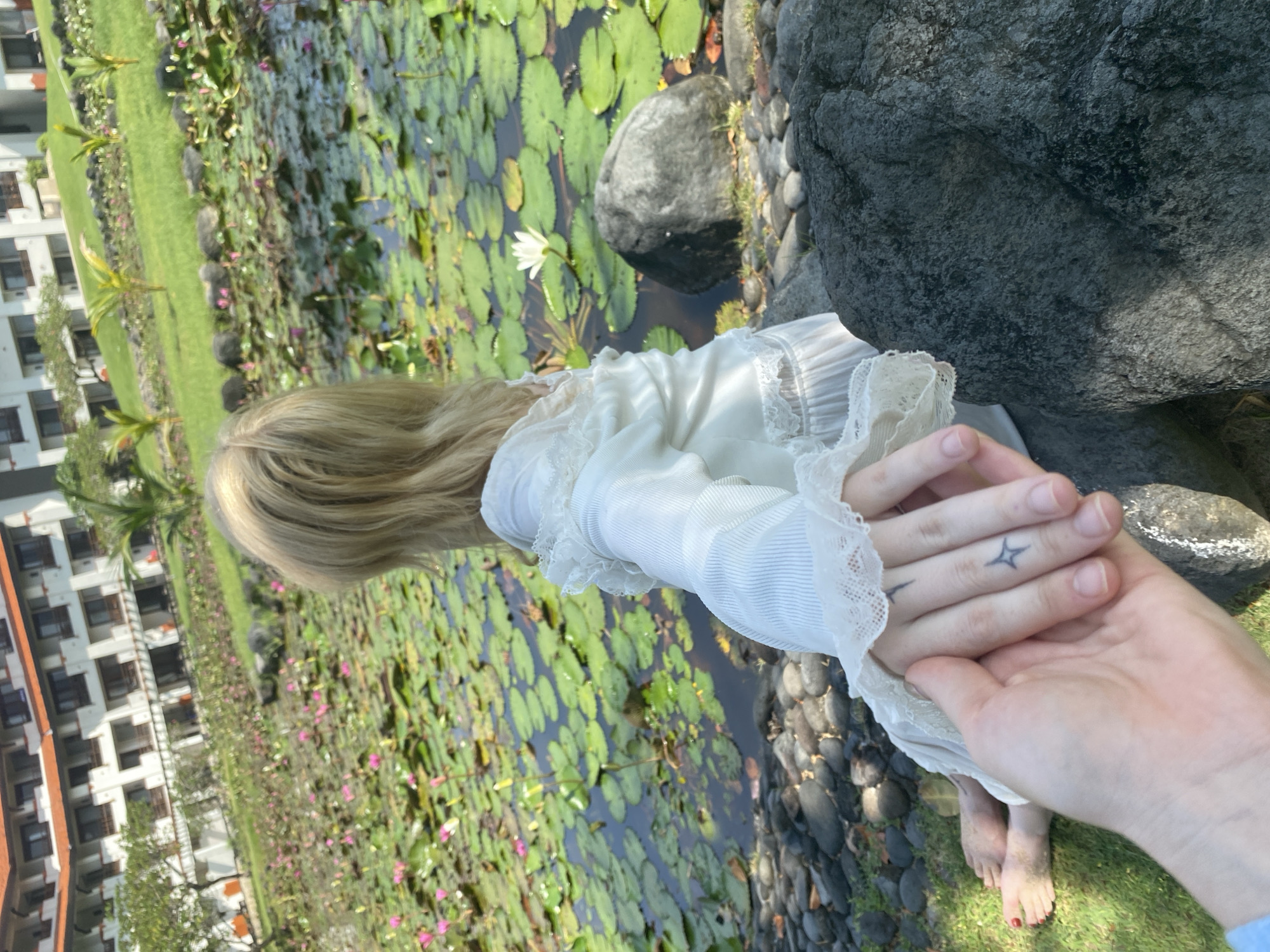 A photo shows a person kneeling by a pond, holding hands with someone. The pond is covered with lily pads and some white water lilies. The kneeling person has blond hair and is wearing a white dress with lace trim. The other person's hand has a tattoo of a four-pointed star. The location appears to be in Nusa Dua, Indonesia, based on the architecture of the buildings in the background.