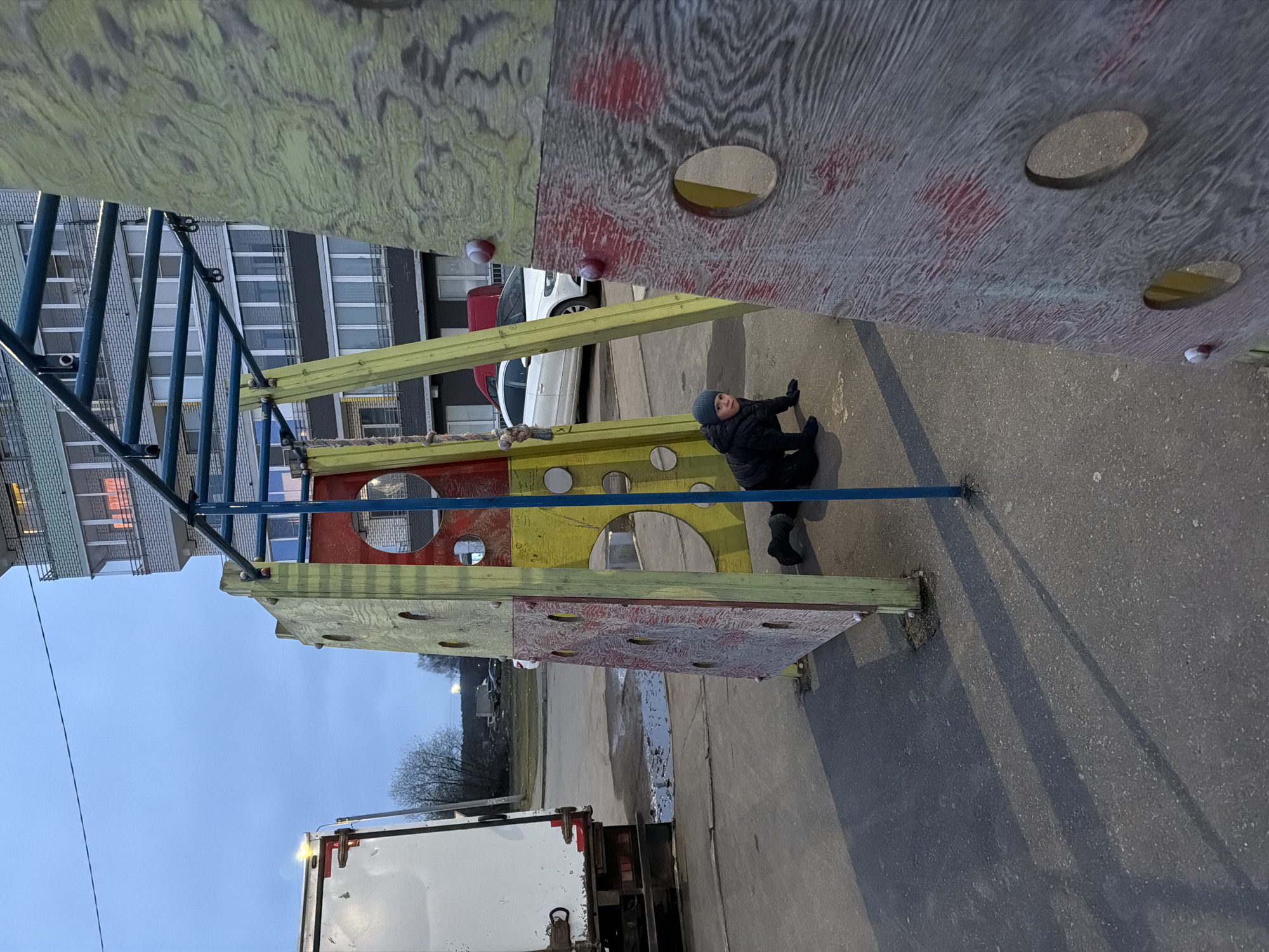 This media file captures an outdoor scene featuring a child near a colorful playground structure in what appears to be an urban or semi-urban setting in Chaikovsky, Russia.
The primary subject is a young child, dressed in a dark winter jacket, a blue knitted hat, and gloves, sitting on the paved ground in the mid-right of the frame. The child is looking towards the camera with a neutral expression.
Dominating the center and right foreground is a multi-part playground structure, constructed from wood panels in yellow, red, and a patterned grey/white. These panels feature numerous circular cutouts, some with contrasting inner colors like yellow or light brown, and some red, circular, knob-like elements, possibly climbing holds. To the left of the wooden structure, a blue metal climbing frame or set of monkey bars is visible.
In the background, several elements indicate an urban environment. A white car and a partially visible red car are parked to the right of the child. Behind them, multi-story buildings with numerous windows stretch across the background. On the far left, the back of a white truck is partially visible. The ground is paved, likely asphalt or concrete, with some shadows cast by the structures. The sky in the upper left is clear or lightly overcast, suggesting daytime, possibly late afternoon, given the soft lighting. Bare trees are visible in the distant background on the left. The child's warm clothing suggests cool or cold weather. There is no visible text.