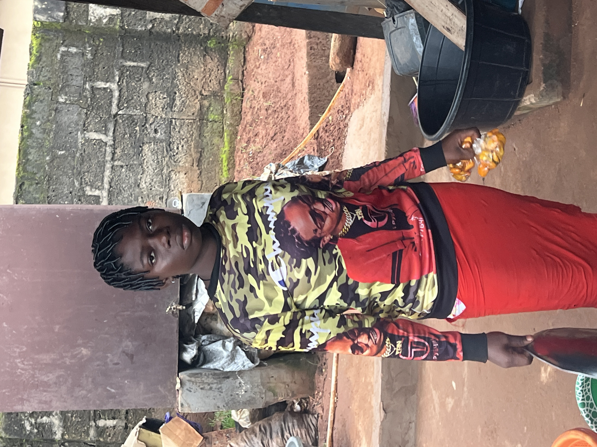 A young Black female with dark braided hair stands in what appears to be an outdoor market setting in Nkpor, Nigeria. She is wearing a camouflage long-sleeved shirt with images of men printed on it and a long, flowing red skirt. In her right hand, she holds two small, clear plastic bags containing what appear to be yellow-orange fruits or candies. Her left hand is holding the handle of a large, flat metal object, possibly a serving tray or a wide knife. To her right, a large black plastic basin is visible, containing a piece of wood. The background consists of a rough-textured, possibly brick wall with patches of moss, and a reddish-brown dirt ground. Other objects, including bags and cardboard boxes, are partially visible in the background, suggesting a commercial or storage area.
