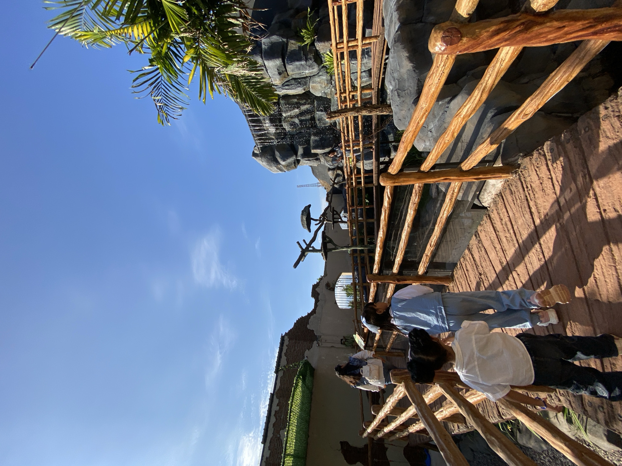 The image depicts a scenic view at Mangli, Indonesia. The scene is composed of a man-made waterfall structure constructed of grey rocks and partially obscured by a wooden fence in the background, and lush green palm fronds at the top of the frame.
In the foreground, two individuals are standing on a brown-colored pathway. Both people are seen from the waist up. One person is wearing a white shirt and dark pants. Next to that person is another individual in a blue denim jumpsuit. Both individuals are facing away from the camera towards the waterfall.
Behind them is a section of a building. The architectural details suggest a historical or rustic design, with decaying brickwork and sections covered in green foliage. Above this building, there is a dark statue on a pole.
The sky is a clear blue with some sparse, light clouds.