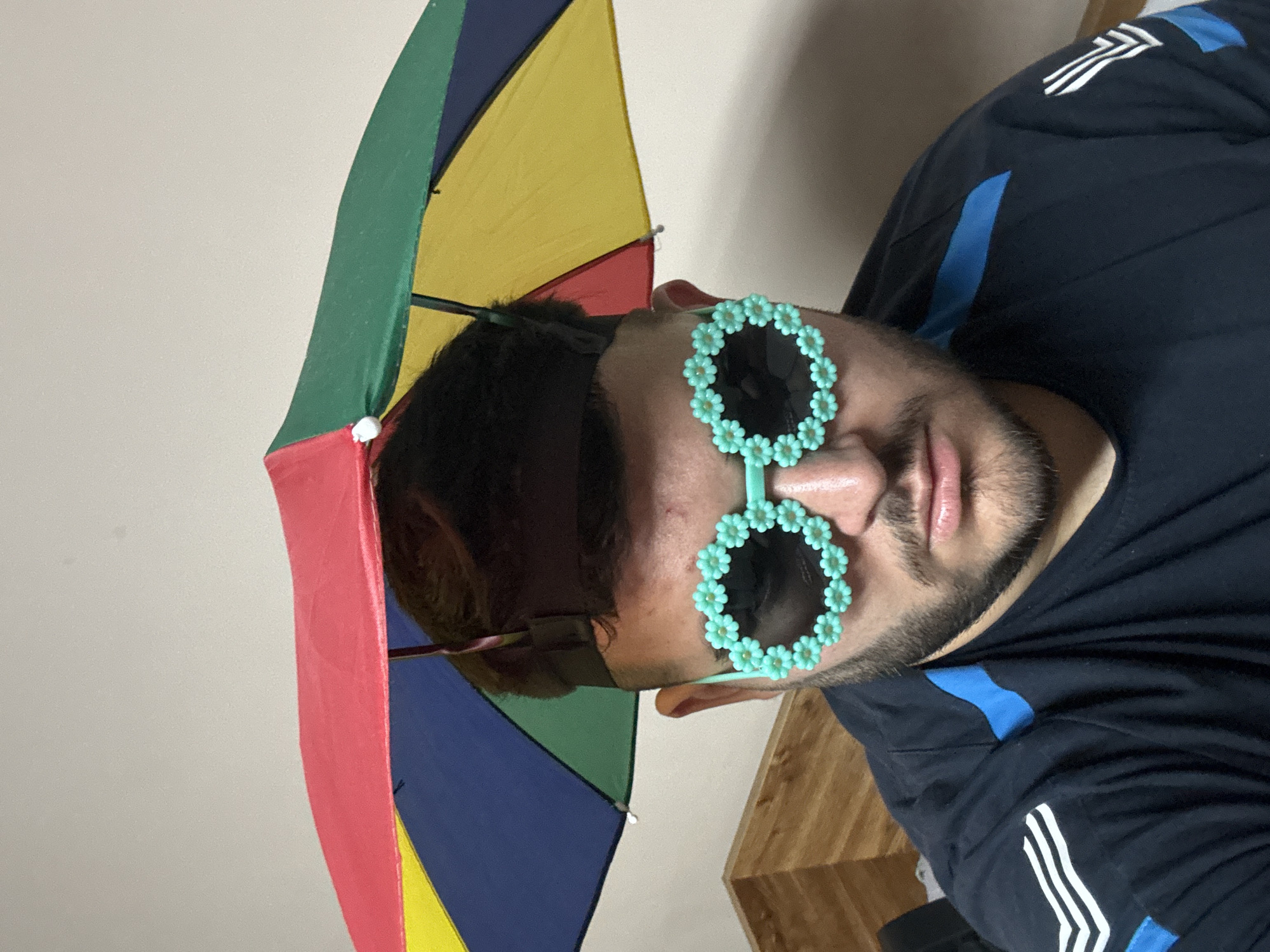 A man with a beard is wearing blue flower sunglasses and holding a small, multicolored umbrella. He is wearing a black shirt with blue and white stripes on the shoulder and sleeve. The background appears to be an interior setting with light-colored walls and wooden furniture. There is no information indicating that the photo was taken in Sofia, Bulgaria.