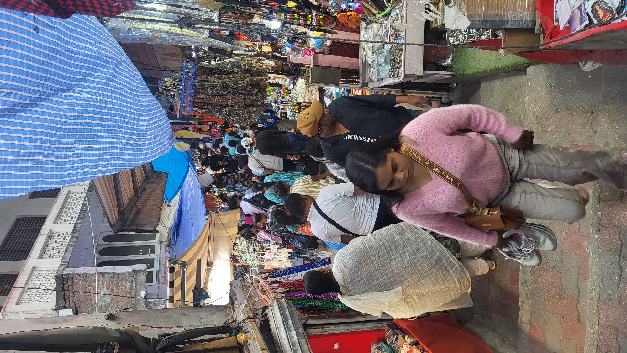 This image captures a bustling street market in Kasol, India, viewed from a slightly elevated perspective looking down a crowded pathway. Numerous people, predominantly adults, fill the narrow aisle between market stalls.
In the foreground, a woman wearing a pink fluffy sweater, light grey pants, and white sneakers with grey laces is prominently featured, looking down to her right. A brown shoulder bag with "Krazy Kat" visible on its strap is slung across her body. Behind her, other shoppers and vendors are densely packed. One man in a black t-shirt and a light brown cap has "LIVE MINDS #" visible on his shirt. Many individuals are seen from behind, engaged in browsing or walking through the market.
The scene is characterized by numerous market stalls lining both sides of the path, displaying a variety of goods such as clothing, accessories, and trinkets. Overhead, multiple tarpaulins and awnings, including a prominent blue and white striped one, a solid blue one, and a beige striped one, provide shade or shelter from the elements. Buildings with windows and patterned grilles are visible behind the awnings. The ground is paved with reddish and grey interlocking bricks or tiles. The overall impression is one of a vibrant, crowded daytime market activity.