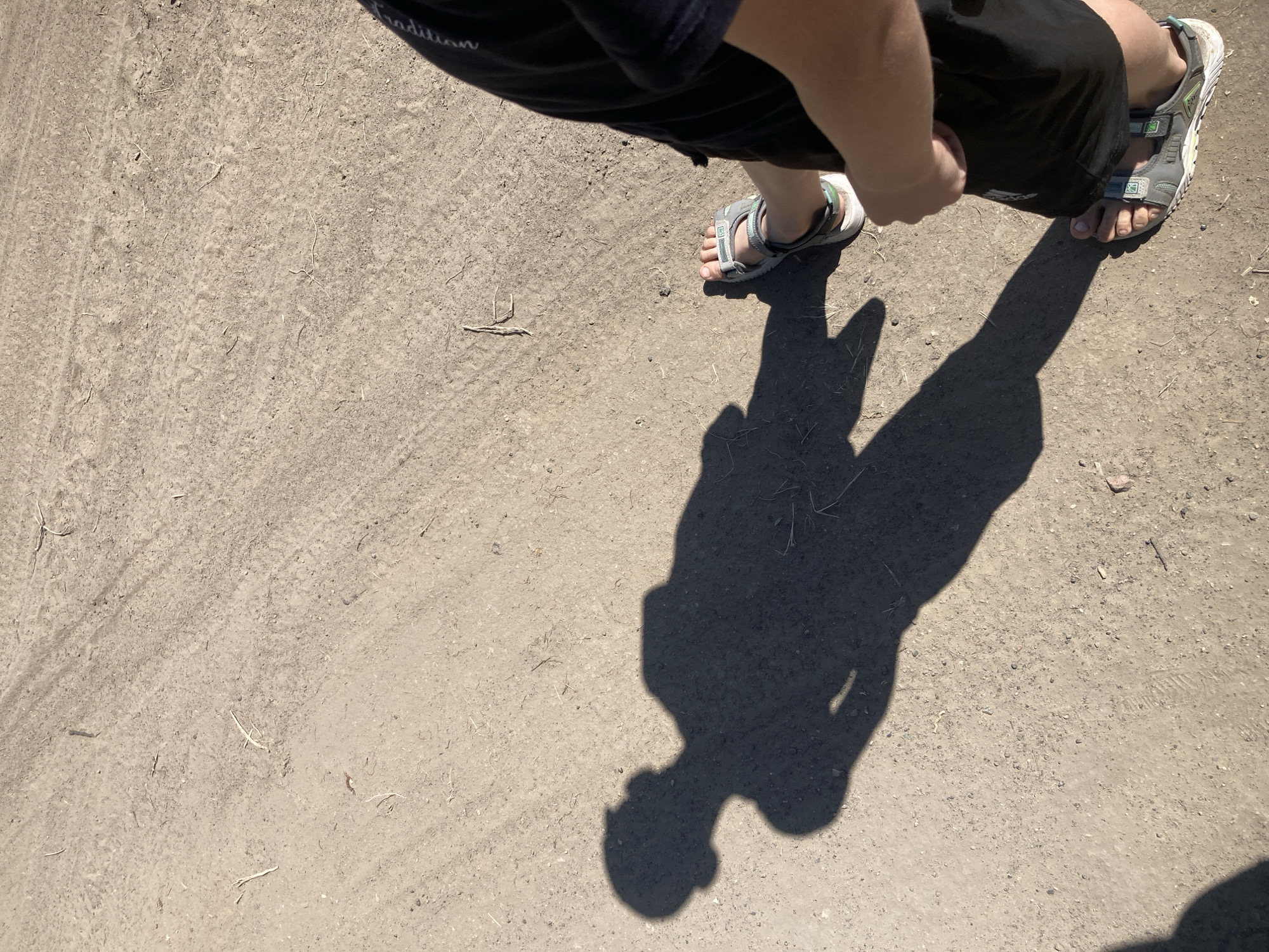 The image shows a person's shadow cast on a dusty, unpaved ground, possibly a rural road or path in Rakity, Russia. The shadow depicts a person with their arm raised, appearing to be walking or standing. The person's feet, wearing sandals, are visible in the lower right portion of the frame. The ground is textured with small stones and dry vegetation. The lighting suggests it is daytime, with the sun casting a long shadow.