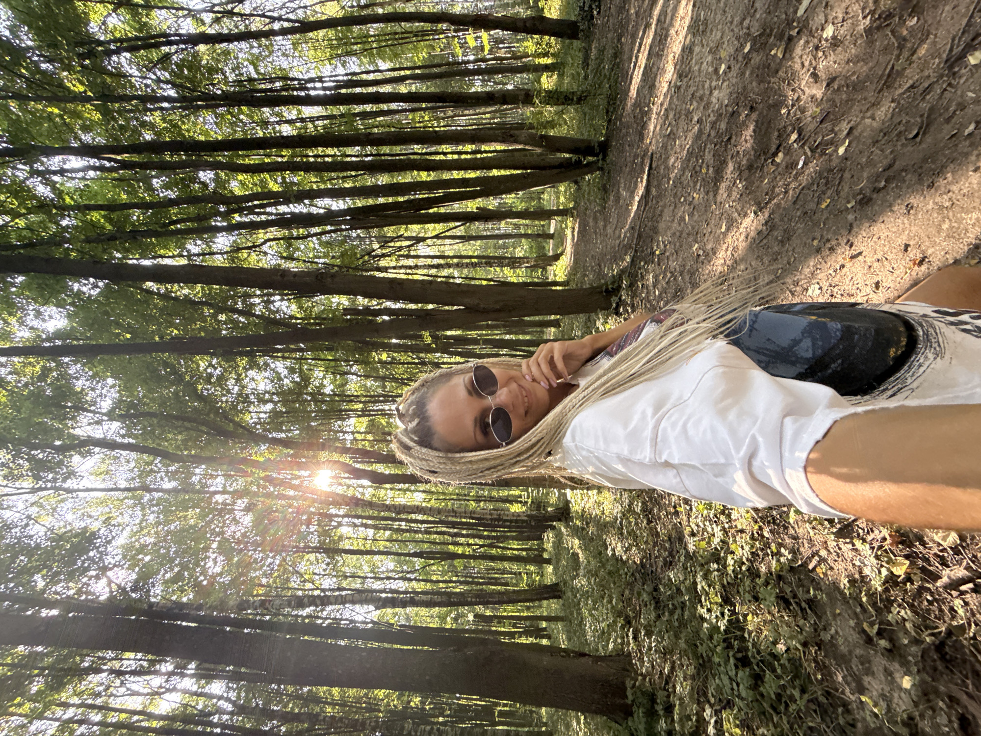 A woman with blonde braided hair and sunglasses is taking a selfie in a sun-dappled forest. She is wearing a white t-shirt and a dark bag is visible over her shoulder. The forest is composed of tall trees with green foliage, and a dirt path winds through the trees in the background. The sun is shining brightly through the leaves, creating lens flares. The ground is covered with leaves and vegetation. The location is identified as Rusanovo, Russia.