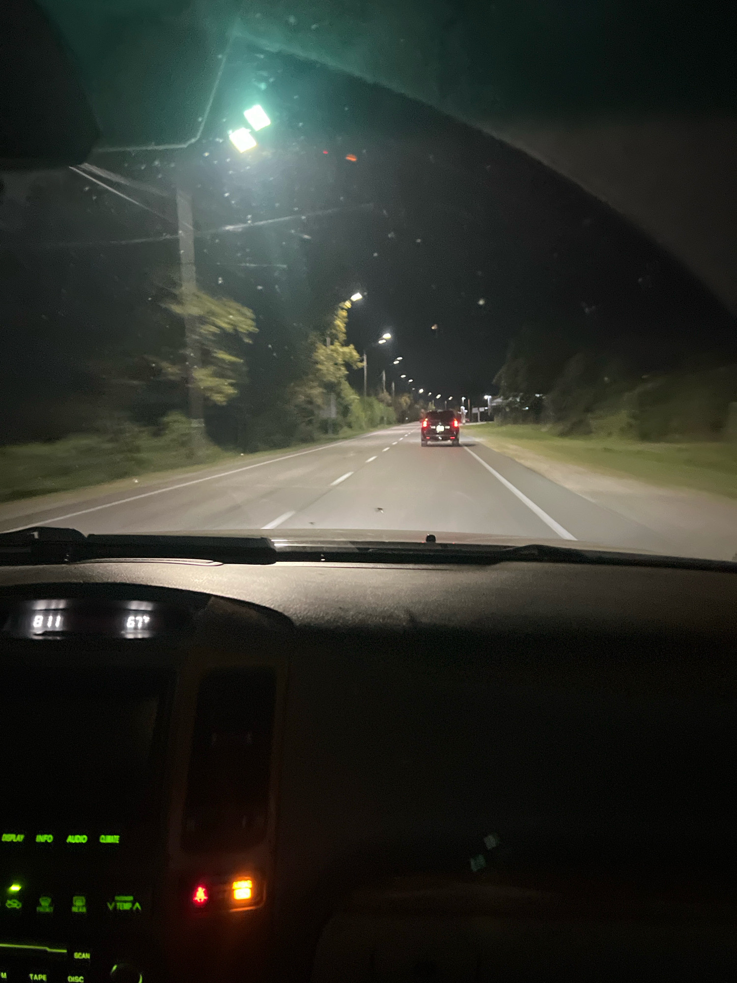 The image depicts a night-time view from inside a car, driving on a road in Primorskoe, Abkhazia. The road is paved and has white lane markings. Streetlights are visible along the roadside, illuminating the scene. Trees and vegetation line the sides of the road. In the distance, another car with its red taillights illuminated is driving in the same direction. The car's dashboard displays the time as 8:11 and the temperature as 67 degrees Fahrenheit. The interior of the car is dark, with some green and orange indicator lights visible on the center console. The overall impression is of a quiet night drive on a suburban or rural road.