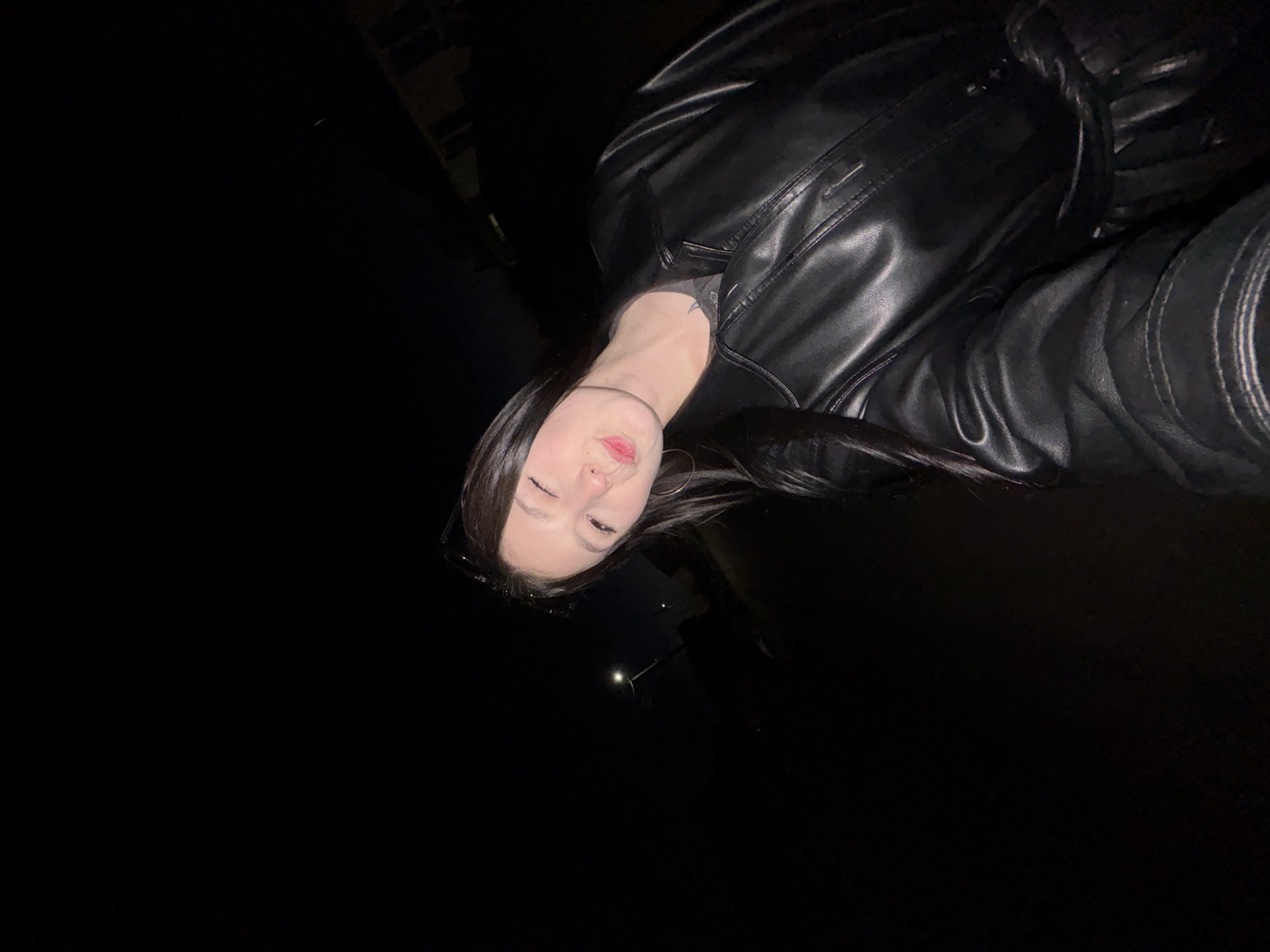 A young woman is wearing a black leather jacket. The location appears to be outdoors in Фоки, Russia, at night, based on the lighting and minimal background details. The woman is facing the camera. There's a street light visible in the far background. The scene is dark.