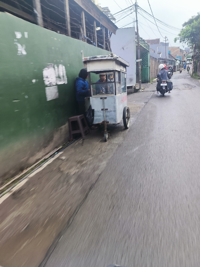 The image shows a street scene in Cimenyan, Indonesia, located at the coordinates -6.888 latitude and 107.652 longitude. A food cart is parked on the side of the street next to a green wall. There are three people present. A person is standing next to the food cart, and two people are seated inside the cart. A motorcycle is parked behind the food cart. The street appears to be paved and is relatively empty. The environment is urban, with buildings and power lines visible in the background. The sky is overcast and the weather appears to be damp.
