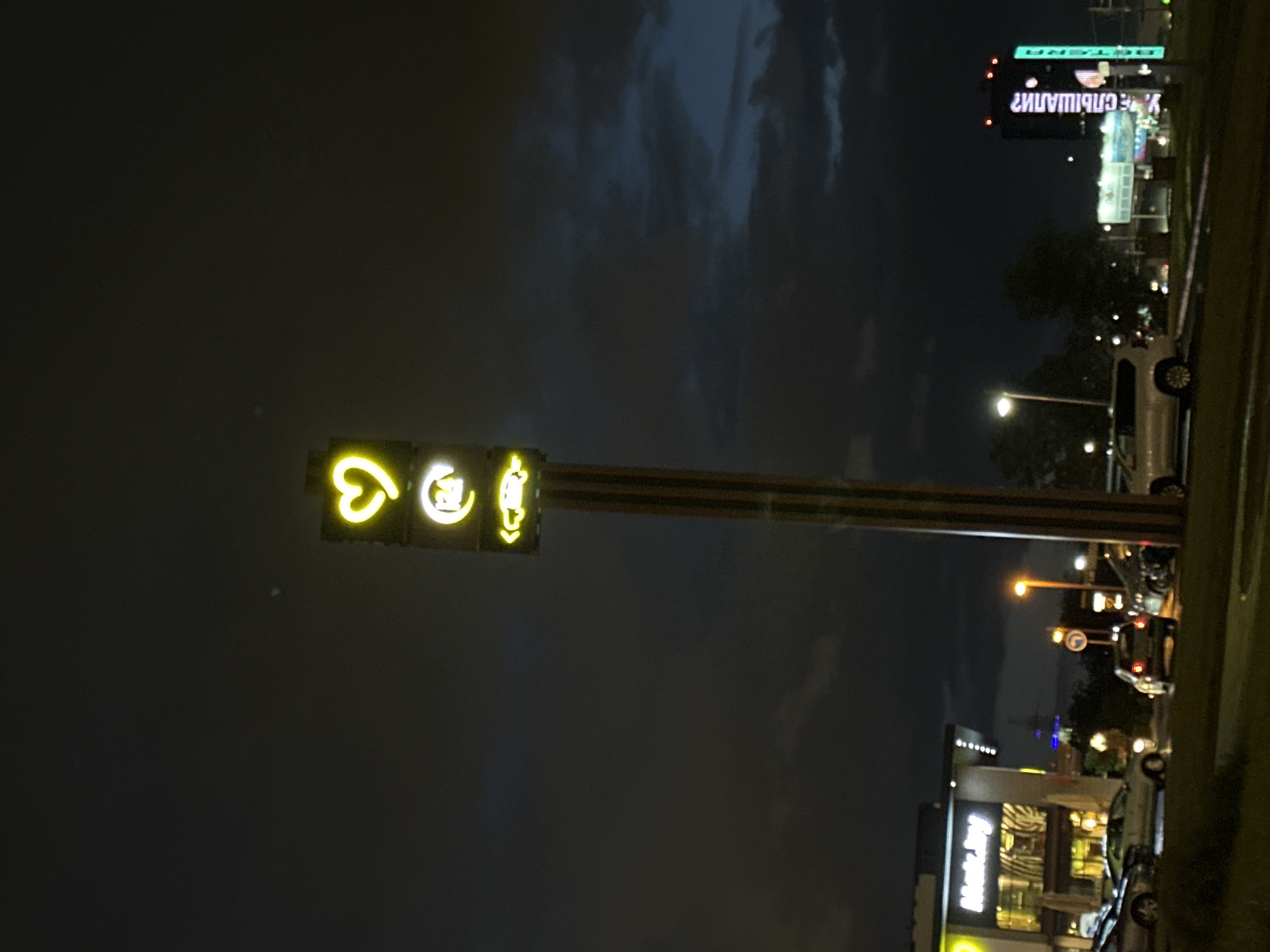 The image depicts a street scene at night in Minsk, Belarus. A brightly lit traffic light with a heart, a plug, and a pedestrian symbol faces left. A tall, narrow building with large, illuminated signs and several streetlights with cars parked on the side of the street are visible. A long, white van is also parked on the right. The sky is dark with patches of lighter clouds. The sign in the top right reads "Е. Свиридова". A shop called "Mart Inn Food" is visible.