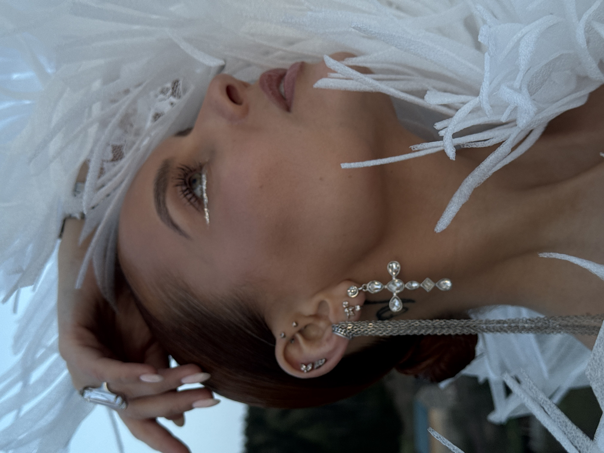 A young woman with red hair pulled back into a bun is shown in a portrait-style image. Her head is turned towards the left, with her gaze directed upwards. She has fair skin, silver eyeliner, and pink lips. She wears multiple silver earrings, including a large cross-shaped earring. She also wears a silver ring on her left hand. She is surrounded by white, feathery material. The background is a blurred blue and green, suggesting an outdoor setting near water. Based on the request, the location is identified as Ольховка, Russia.