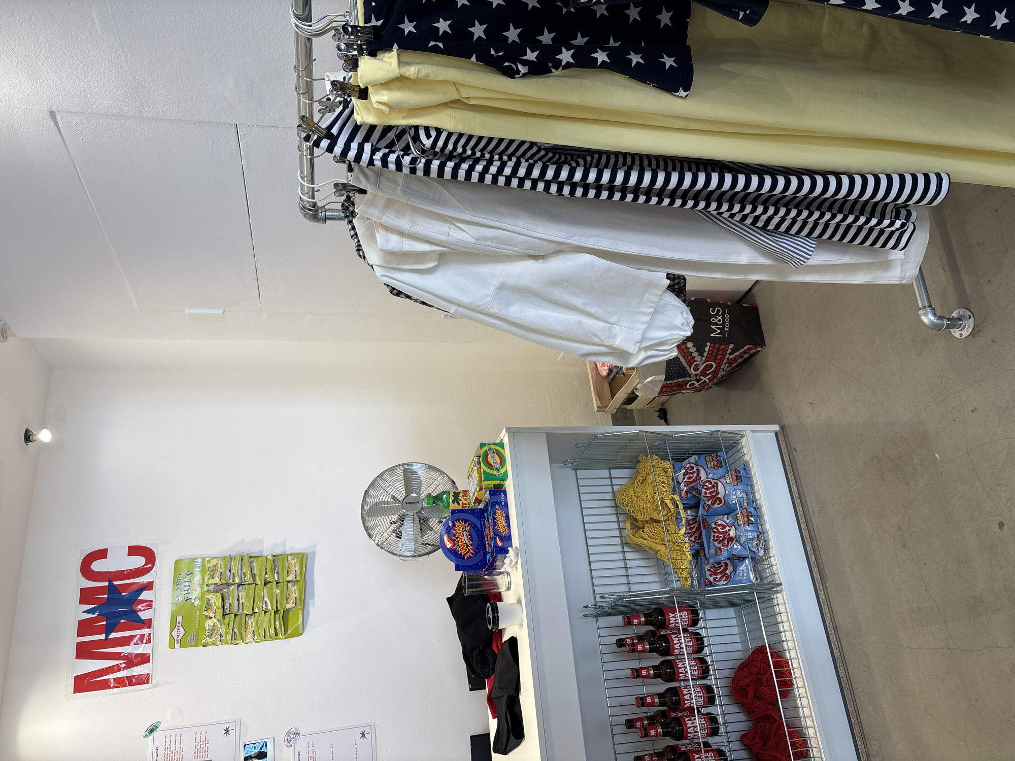 The image depicts the interior of a clothing store located in London, United Kingdom.
In the upper portion of the frame, clothes hang on a metal rack. The garments include items with star patterns, yellow fabric, black and white stripes, and white fabric. Below this, near the center, is a cooler filled with items like potato chips, beer, and red bandanas. A fan sits beside the cooler. On the wall to the left, there are posters including one with "MMC" emblazoned on it and another with "SCAMPI FRIES". Also present is text related to a clothing store and prices. Further in the background, there is a cardboard box labeled "M&S - FOOD". The floor is concrete.