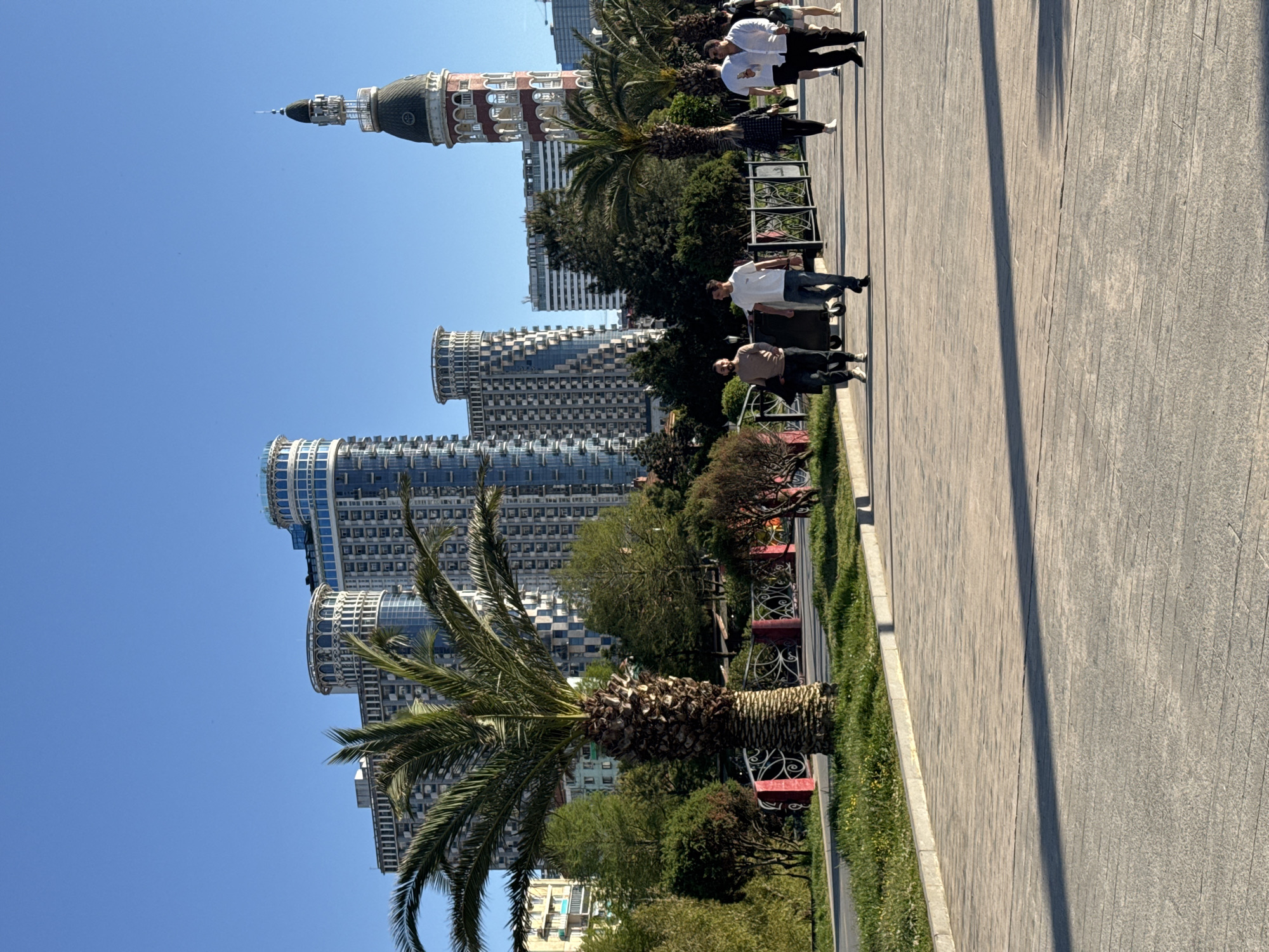 The photograph captures a scene in Batumi, Georgia, showcasing a blend of modern architecture and pedestrian activity on a sunny day. In the background, several high-rise buildings with rounded facades are visible, suggesting a coastal or waterfront development area. To the right of these buildings, a more traditional structure with a distinctive dome and tower is present.
A paved walkway occupies the right side of the image, where several people are walking or standing. The people are dressed in casual clothing. Adjacent to the walkway is a landscaped area with trees, bushes, and small red-trimmed fences, adding a touch of greenery to the urban setting. The presence of palm trees in the foreground further emphasizes the coastal location of the city. The lighting is bright and clear, allowing for sharp details and distinct shadows to be observed.