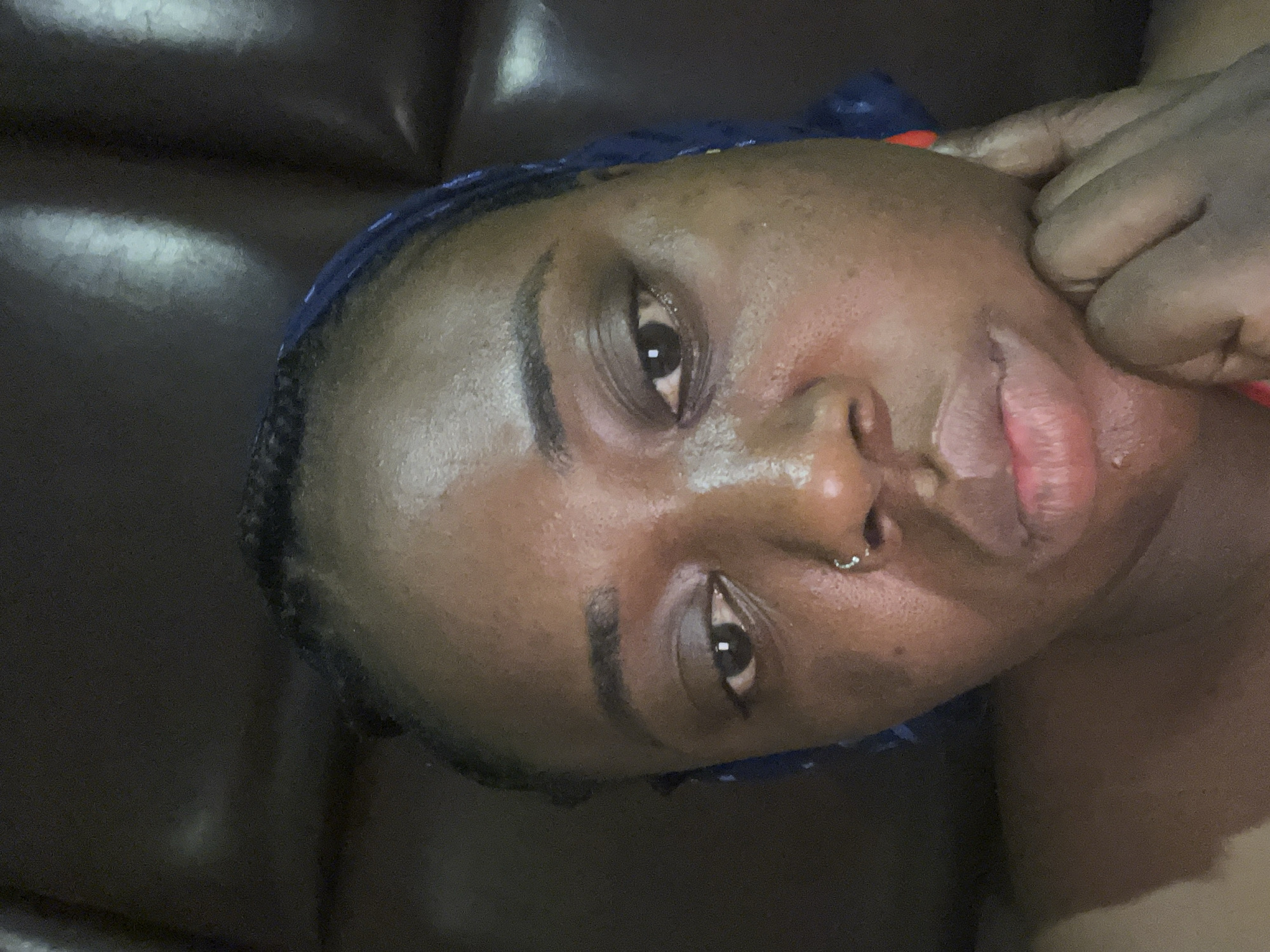 The image shows a close-up of a dark-skinned person's face. They have a nose ring and are wearing a blue head covering. Their hand is near their face, possibly touching it. The background appears to be a dark, textured surface, potentially a leather couch. There is no information in the image to identify the location as Abuja, Nigeria.