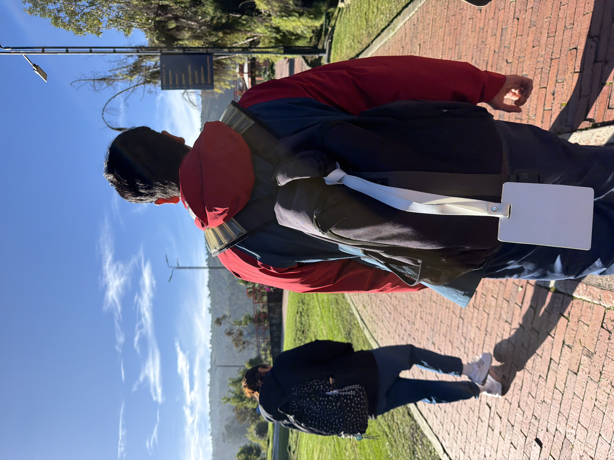 An outdoor scene features two individuals walking away from the camera on a brick pathway. The person in the foreground, wearing a red and blue jacket and a black backpack with a white tag, is walking to the right. The person in the background, wearing a black jacket and a black and white patterned backpack, is walking to the left. They are in a park-like setting with green grass, trees, and a clear blue sky with scattered clouds. A lamppost and a sign are visible to the right. The location is identified as Bojacá, Colombia.