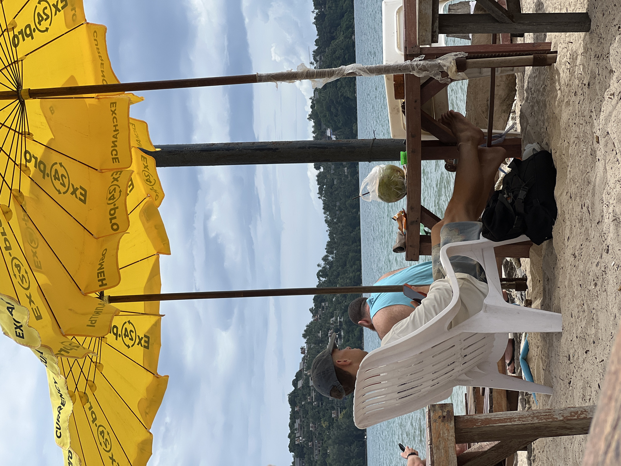 The image depicts a beach scene with two men seated at a wooden table under yellow umbrellas. In the foreground, a man wearing a baseball cap and a light-colored shirt is seated in a white plastic chair, facing away from the camera, with his legs propped up on a horizontal bar. He is holding a smartphone in his right hand. Beside him, another man in a blue tank top is also seated, and a coconut in a plastic bag rests on the table. The background shows a body of water, a lush green hillside covered with houses, and a cloudy sky. Several yellow beach umbrellas with text printed on them are visible in the upper portion of the image. The ground is covered with sand.
