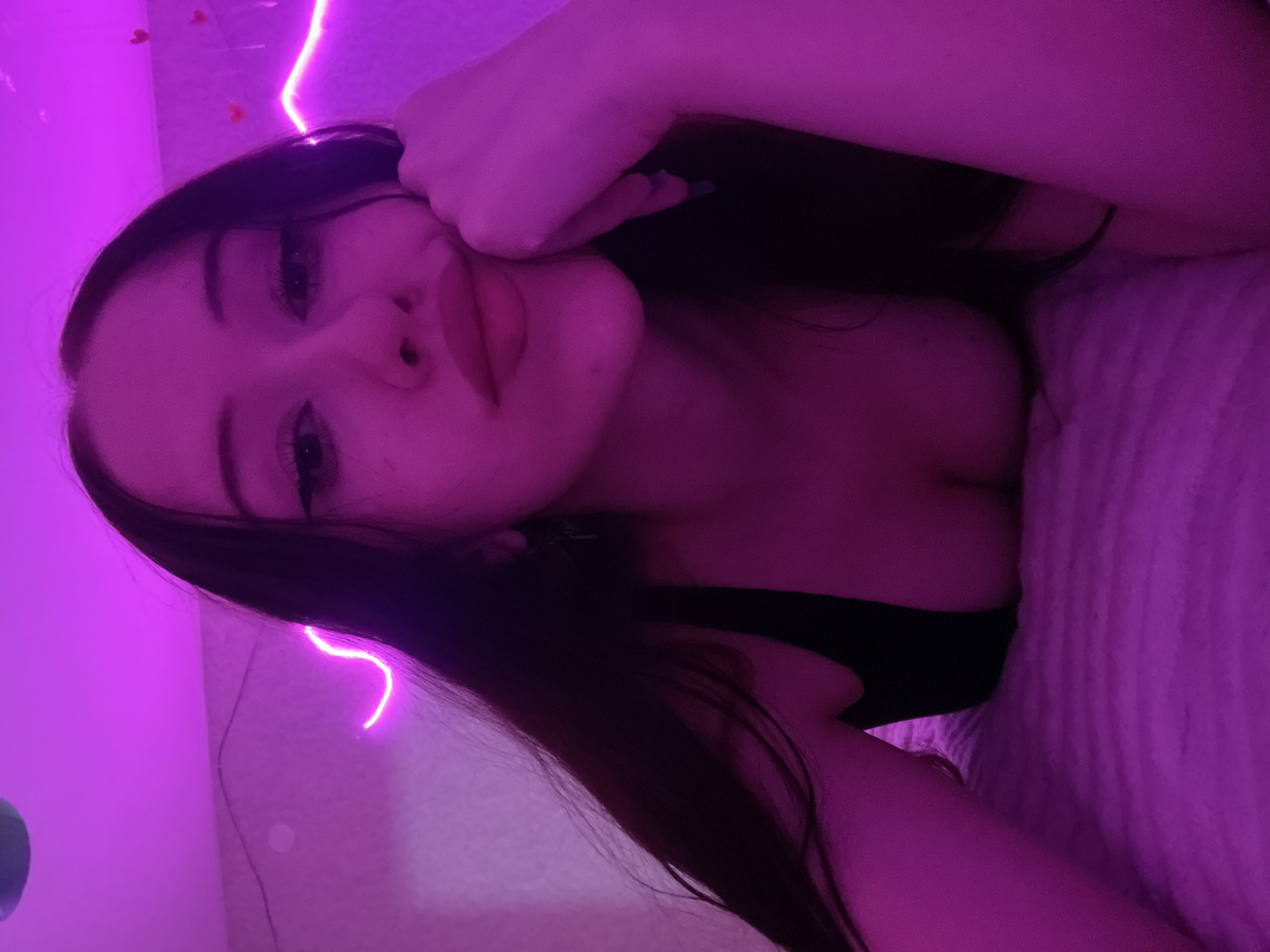 A young woman with long, dark hair is lying on a bed in a room illuminated by purple neon lights. She is looking towards the camera with her right hand resting on her cheek, and her lips are slightly parted. She is wearing a black top. The room appears to be a bedroom, with a textured blanket on the bed and heart-shaped decorations on the wall behind her, next to the neon lights.