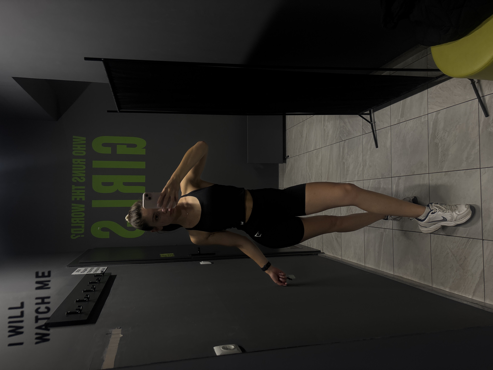 A young woman in athletic wear takes a mirror selfie in what appears to be a gym locker room. She is wearing a black sports bra and black shorts, with white Nike sneakers. The walls are dark gray, with motivational text on them, one reading "I WILL WATCH ME" and another, mirrored, "WHO RUNS THE WORLD? GIRLS." The floor is tiled in a light gray marble pattern. A black screen is set up in the corner of the room. A yellow stool is partially visible in the lower right corner. The image is a personal photograph taken by the subject, likely in Varna, Bulgaria, given the context provided.