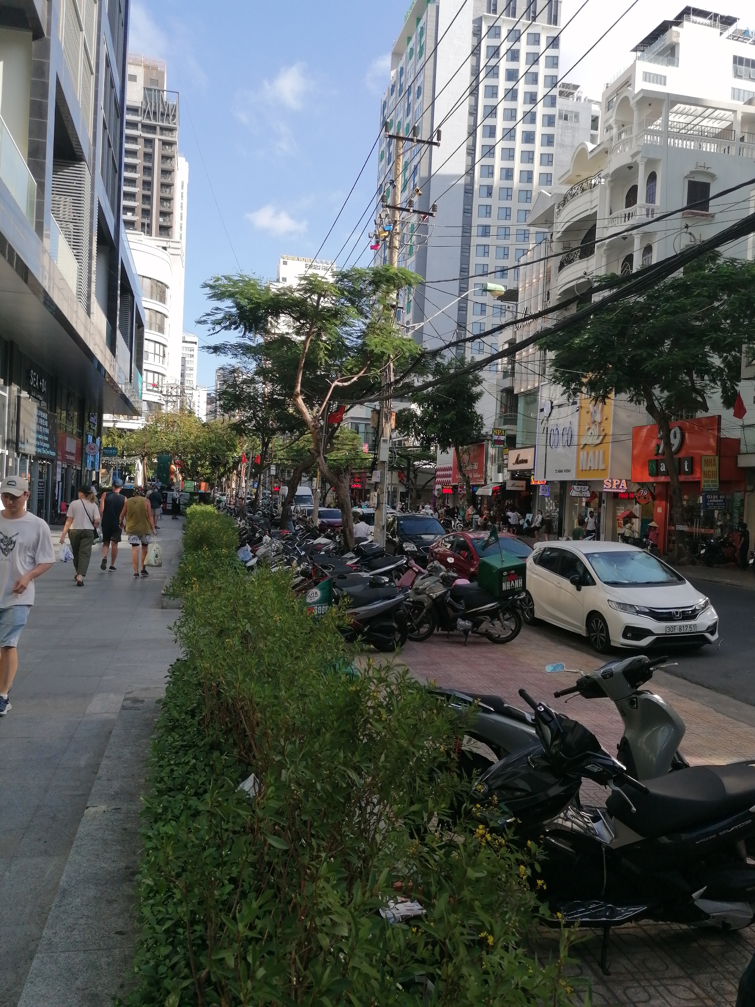 The image captures a street in Nha Trang Ward, Vietnam. On the left is a wide sidewalk with pedestrians walking, lined with a hedge. Parked motorcycles are a prominent feature, lining the sidewalk and the street. A white car is parked on the street, registered 30F-817.51. The street is framed by buildings of varying heights, including modern glass structures and older architectural styles. Overhead, electrical wires intersect the sky. Commercial signage is visible on the buildings, indicating a mix of businesses, including spas, nail salons, and restaurants. The sky is partly cloudy, suggesting a sunny day.