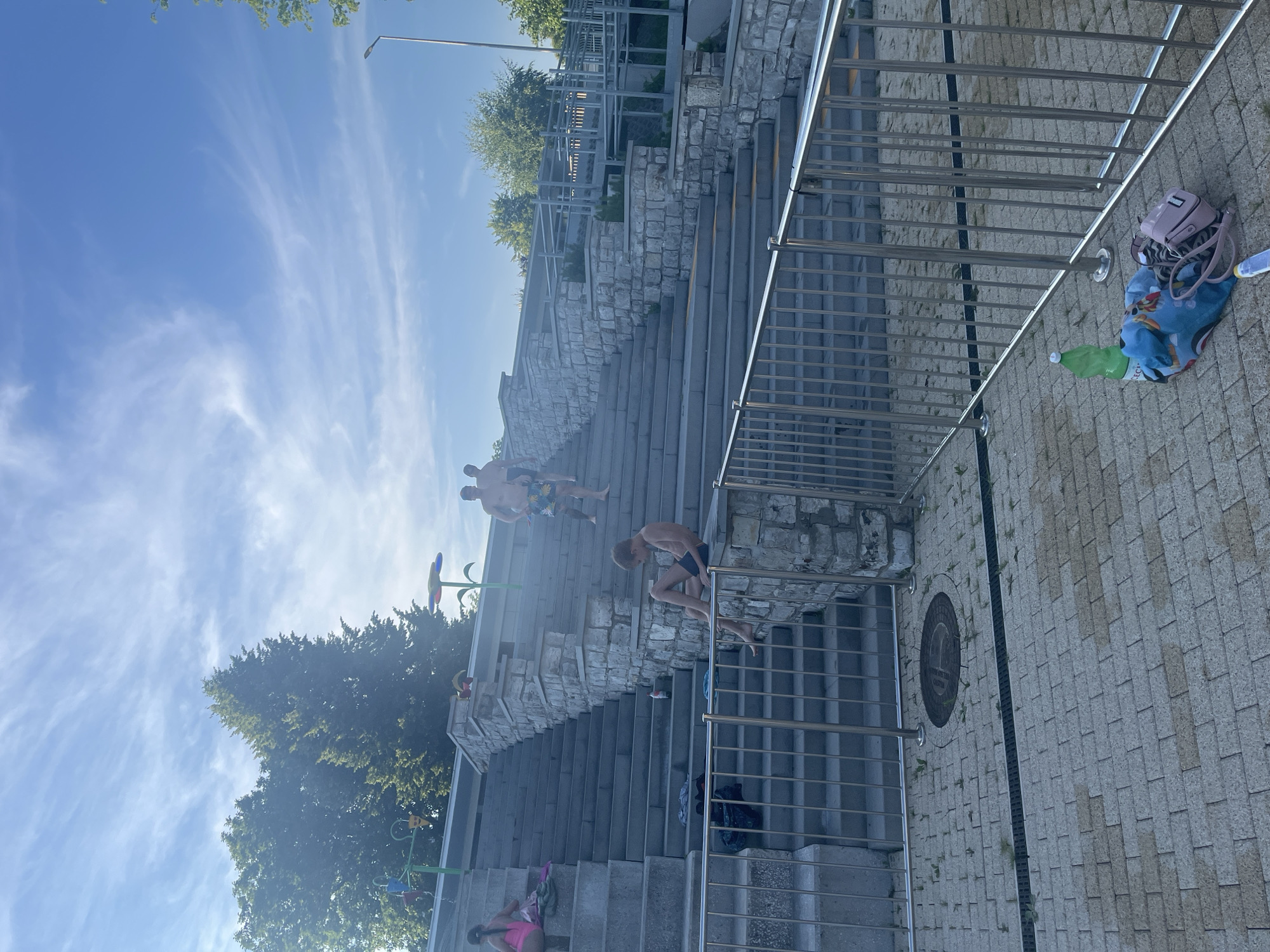 The image depicts an outdoor scene with a focus on tiered stone stairs and a paved area. There are several individuals visible on the stairs, including a young boy sitting on a ledge, appearing to be resting. Further up the stairs, two men in swim trunks are walking. On the left side of the stairs, a young girl in a pink swimsuit is also seated. Various items are scattered on the steps and the paved area, such as towels, bags, a bottle of water, and a pink purse. The environment includes trees and a clear blue sky with wispy clouds, suggesting a warm, sunny day. Metal railings are present along the paved area and the stairs, indicating a public space, possibly a park or a recreational area near water. A manhole cover is visible on the paved ground.