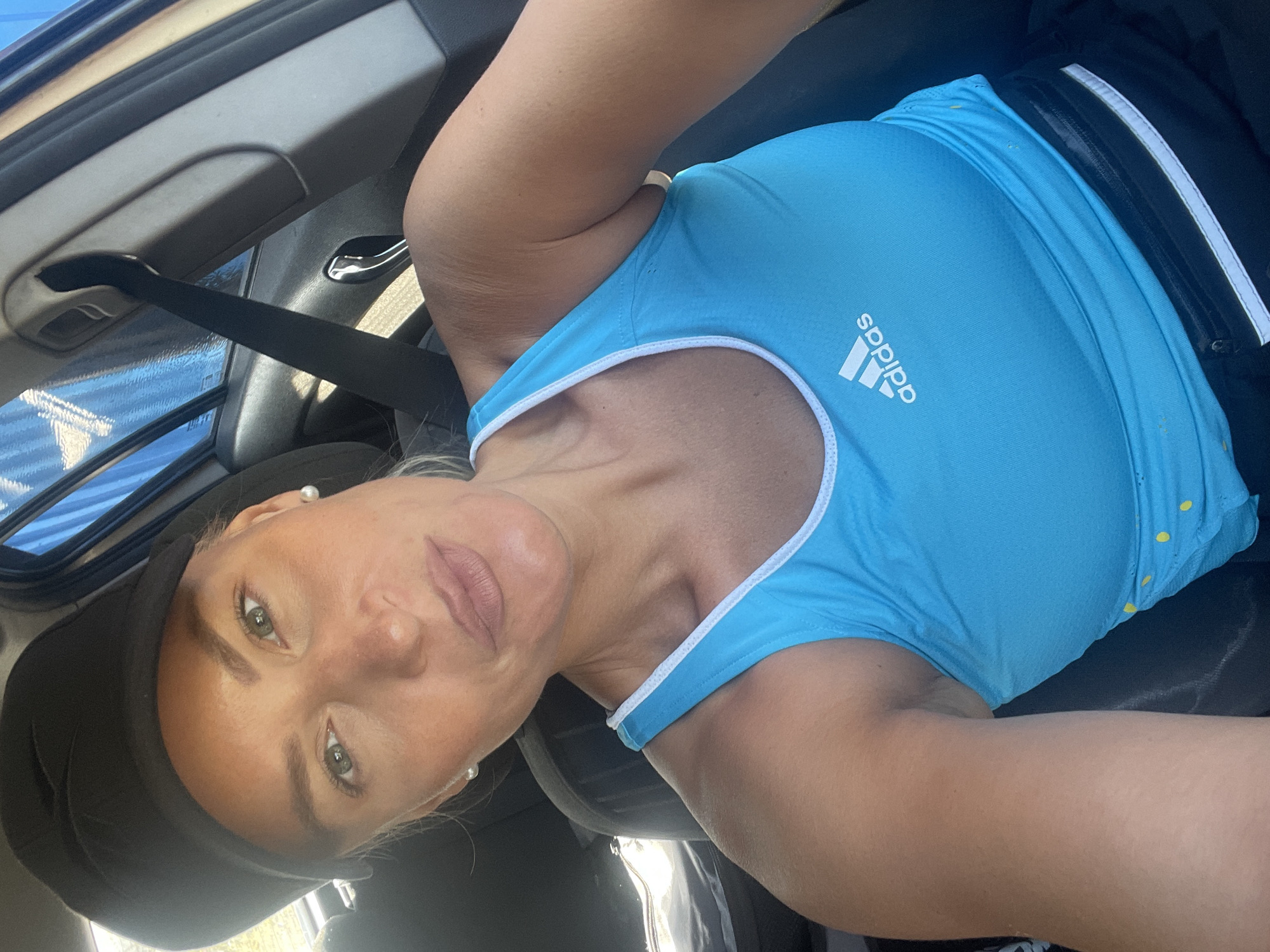 The image is a close-up, selfie-style photograph of a blonde woman wearing a black baseball cap and a bright blue Adidas tank top with white trim. She has light-colored eyes, visible makeup, and is wearing pearl earrings. The woman is sitting in the driver's seat of a car. Visible in the background are a car's interior elements, including a seatbelt, door handle, and part of the window with a blue tarp or curtain visible outside. The location is specified as Baranovka, Russia.