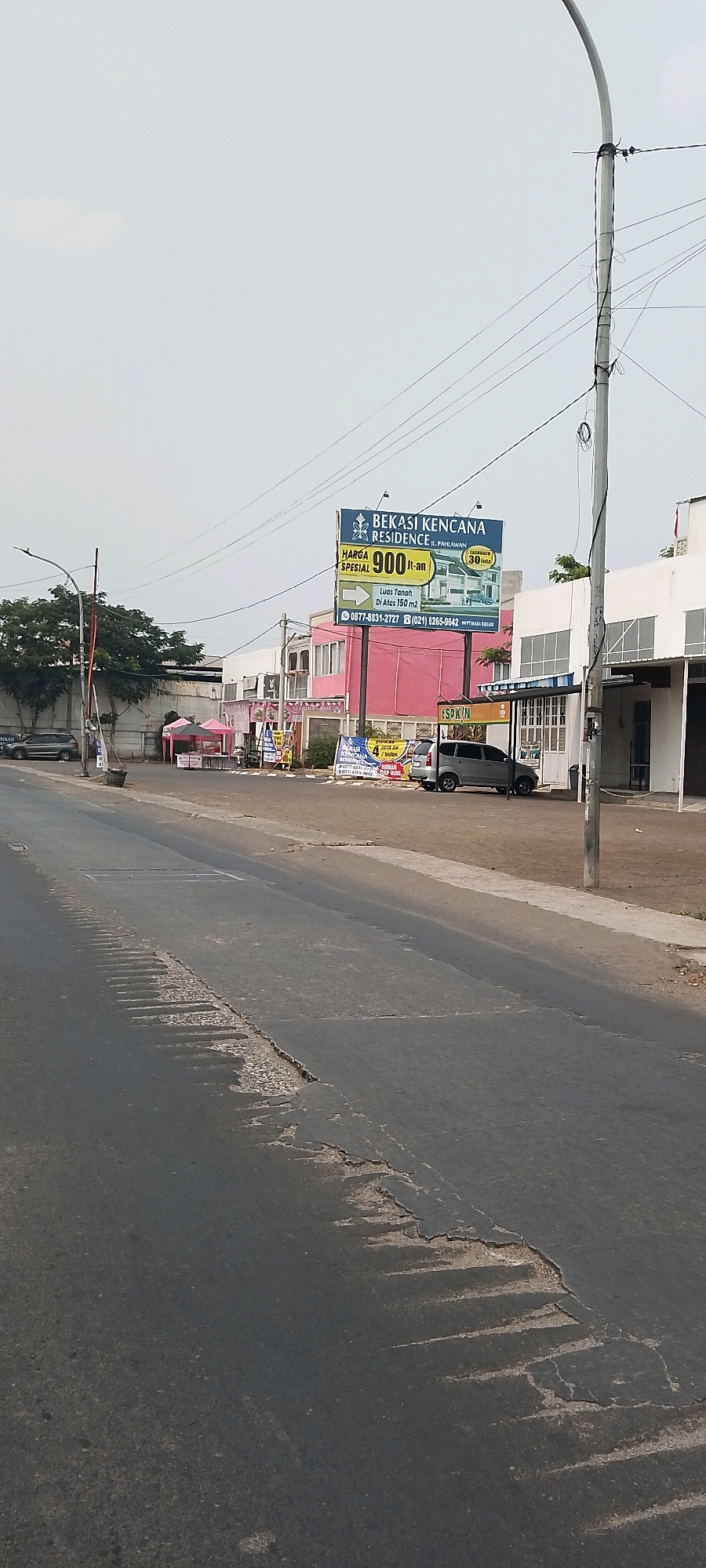 The image depicts a street scene in Bekasi, Indonesia, characterized by a moderately wide asphalt road with visible wear and tear, including cracks and patches. On the right side of the road, a modern building with white and gray facades is present. To the left, a billboard advertising "Bekasi Kencana Residence" with a special price of 900 million Indonesian Rupiah is prominently displayed. Next to the billboard, a pink building with large windows is visible, and further to the left, there are several tents set up, possibly for a market or event. A dark-colored van and a silver SUV are parked on the side of the road. Overhead, power lines are strung between poles, and the sky is overcast. The overall environment suggests a commercial or residential area undergoing development or with active commerce.