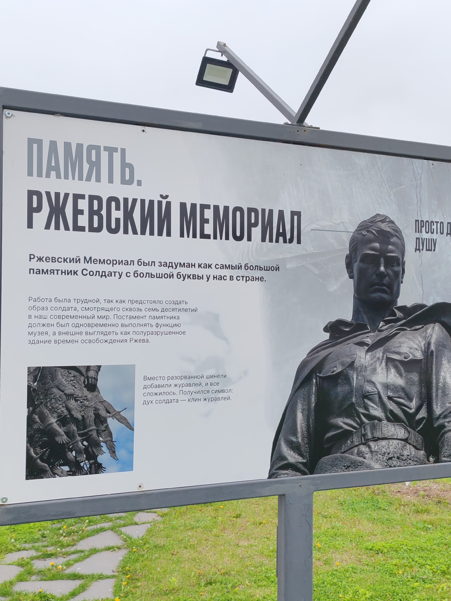 This is a close-up shot of a sign promoting the Rzhev Memorial, located in the city of Tver, Russia. The sign has large lettering announcing "MEMORY. RZHEV MEMORIAL". Beneath this title, there is a description of the memorial as the largest monument to a soldier in the country. It explains the work involved in creating the image of a soldier looking through the decades into the modern world. The text specifies that the pedestal of the monument should simultaneously function as a museum and look like a partially destroyed building from the time of the liberation of Rzhev. The sign also mentions that instead of a torn overcoat, cranes were added, which turned out to be a symbol of the soldier's spirit - a wedge of cranes. Next to the description is an image of the statue, which depicts the upper half of a soldier in uniform, as well as the group of crane statues. The sign is displayed on a grey metal frame. In the background, there is green grass.