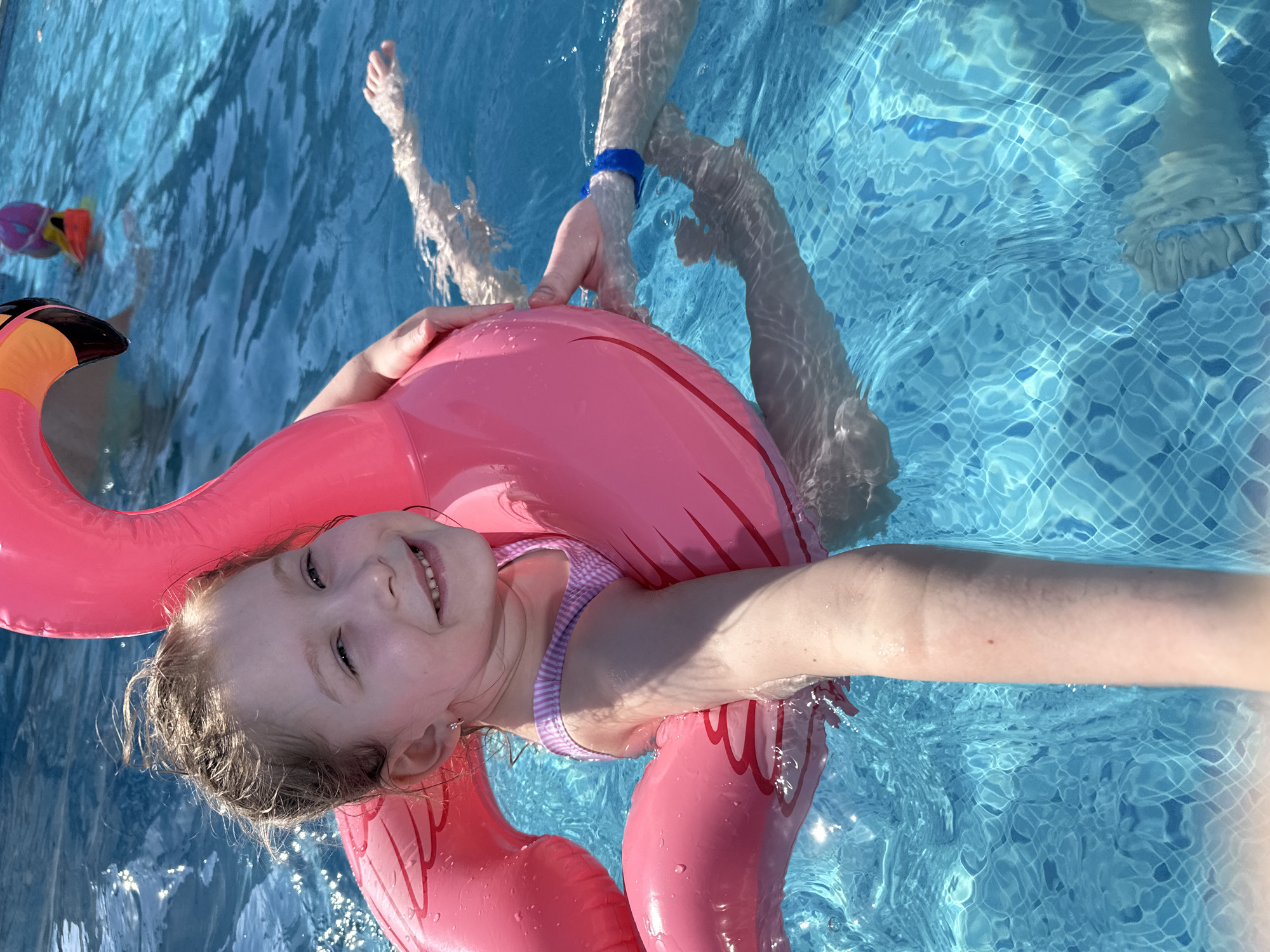 A young child in a striped swimsuit floats in a swimming pool, supported by an inflatable pink flamingo. The child is smiling. Another person's hands are visible on the inflatable flamingo, and other people's legs and feet are also visible in the pool. The bottom of the pool is tiled. The location cannot be determined from the image.