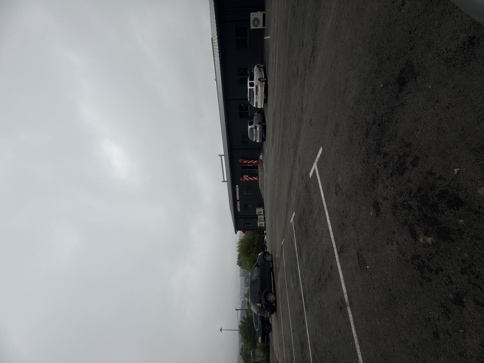 The image depicts a parking lot in front of a commercial building. The building is dark gray with a silver roof and features a sign with Russian text. Several cars are parked in the lot, including a black SUV, a silver hatchback, and a white station wagon. There are also air conditioning units visible on the exterior of the building. The sky is overcast, and the overall scene appears to be in an urban or suburban setting, possibly in Vladivostok, Russia, given the Russian signage. The parking lot is paved and marked with white lines. In the background, trees and distant buildings are visible.