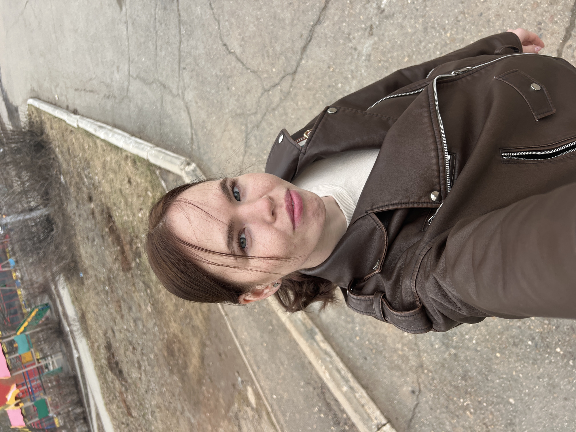 A medium, low-angle shot of a young woman with light skin, brown hair, and blue eyes, facing the camera. She wears a brown leather jacket over a light-colored top.
The background consists of a concrete ground surface with cracks and patches. A strip of curb borders a patch of bare earth with some sparse vegetation. Further in the background, indistinct elements suggest a children's playground with some colorful equipment, indicating an outdoor space in the city of Chaikovsky, Russia.