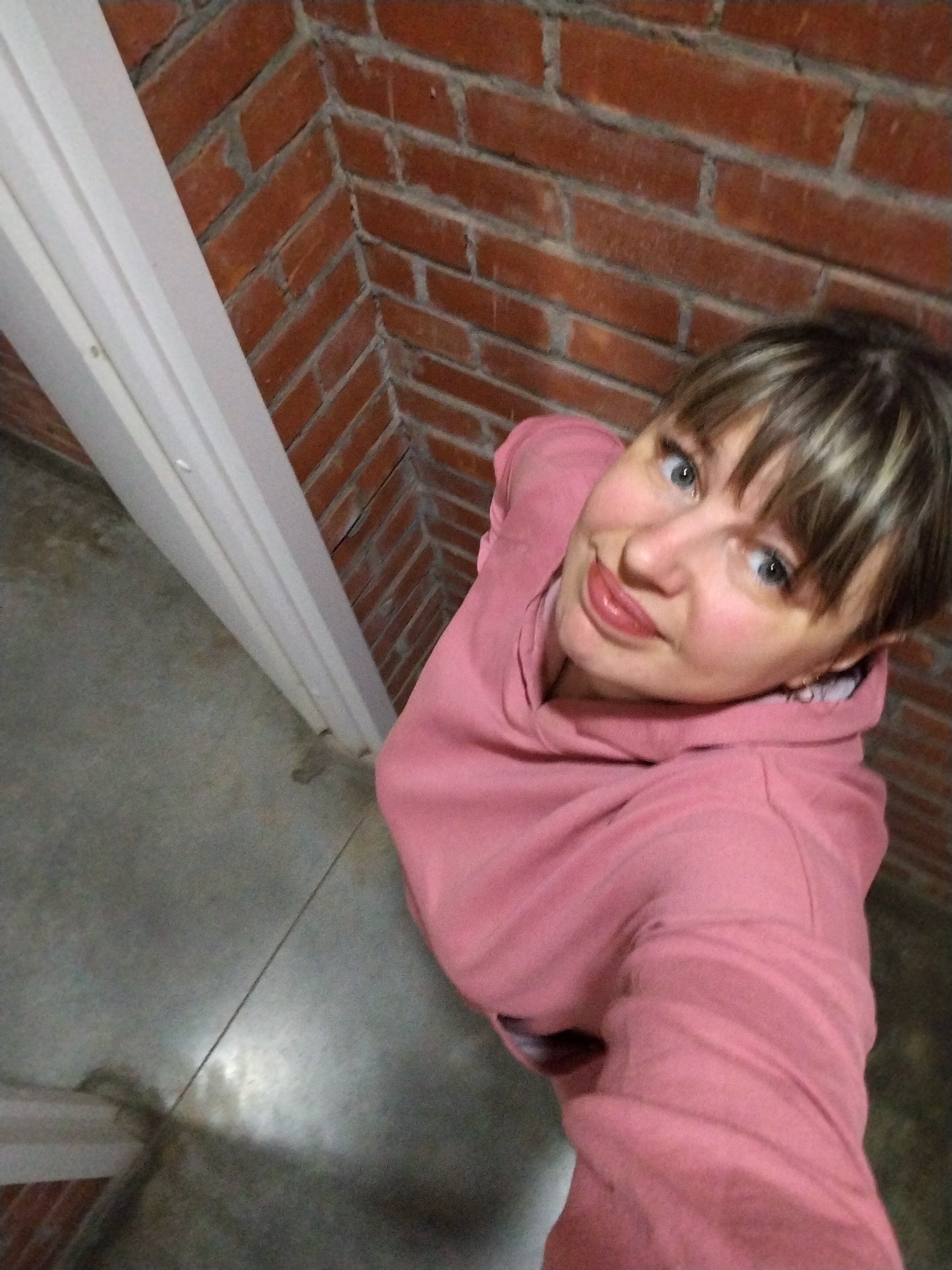 A woman is featured prominently in a high-angle shot, looking up at the camera with a slight, friendly smile. She has medium-length, light brown hair with blonde highlights and bangs. Her eyes are a light blue-green, and her lips are adorned with a natural pink lipstick. She is wearing a casual, solid pink hooded sweatshirt, which appears comfortable and soft.
The setting is an indoor space, characterized by a distinct red-brown brick wall that forms a sharp corner behind her. The bricks are laid in horizontal courses with light-colored mortar. To the left side of the frame, a white vertical structure, possibly a pillar, door frame, or trim, runs diagonally, contrasting with the brickwork. The floor is a muted gray, appearing to be concrete or stone, showing some subtle texture and shading.
The woman's upward gaze and direct engagement with the camera suggest she is posing for a photo, possibly a selfie. Her expression conveys a sense of pleasantness and approachability. There are no other individuals or specific objects identifiable in the scene beyond the architectural elements. The lighting appears to be ambient indoor light, and the time of day cannot be determined. The combination of brick and concrete gives the space a somewhat rustic or industrial-chic aesthetic. This scene is located in Rusanovo, Russia. There is no visible text in the image.