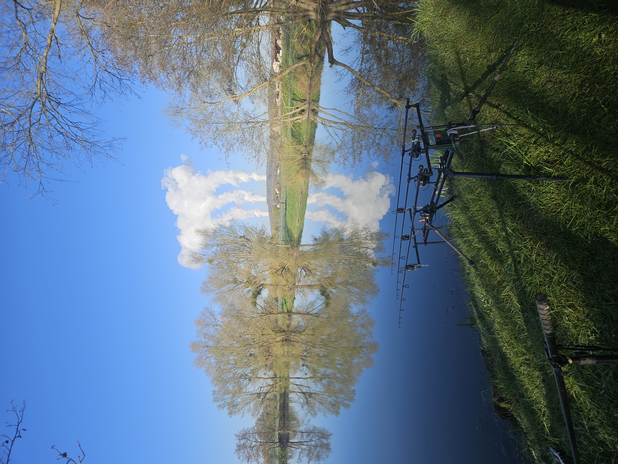A low-angle perspective of a body of water (likely a river or lake) reflects the sky, trees, and land, creating a symmetrical composition. Fishing rods are set up on the right bank, standing in green grass. The reflected trees, the sky, and clouds create an abstract mirror image in the water. The location is likely Gavisse, France.
